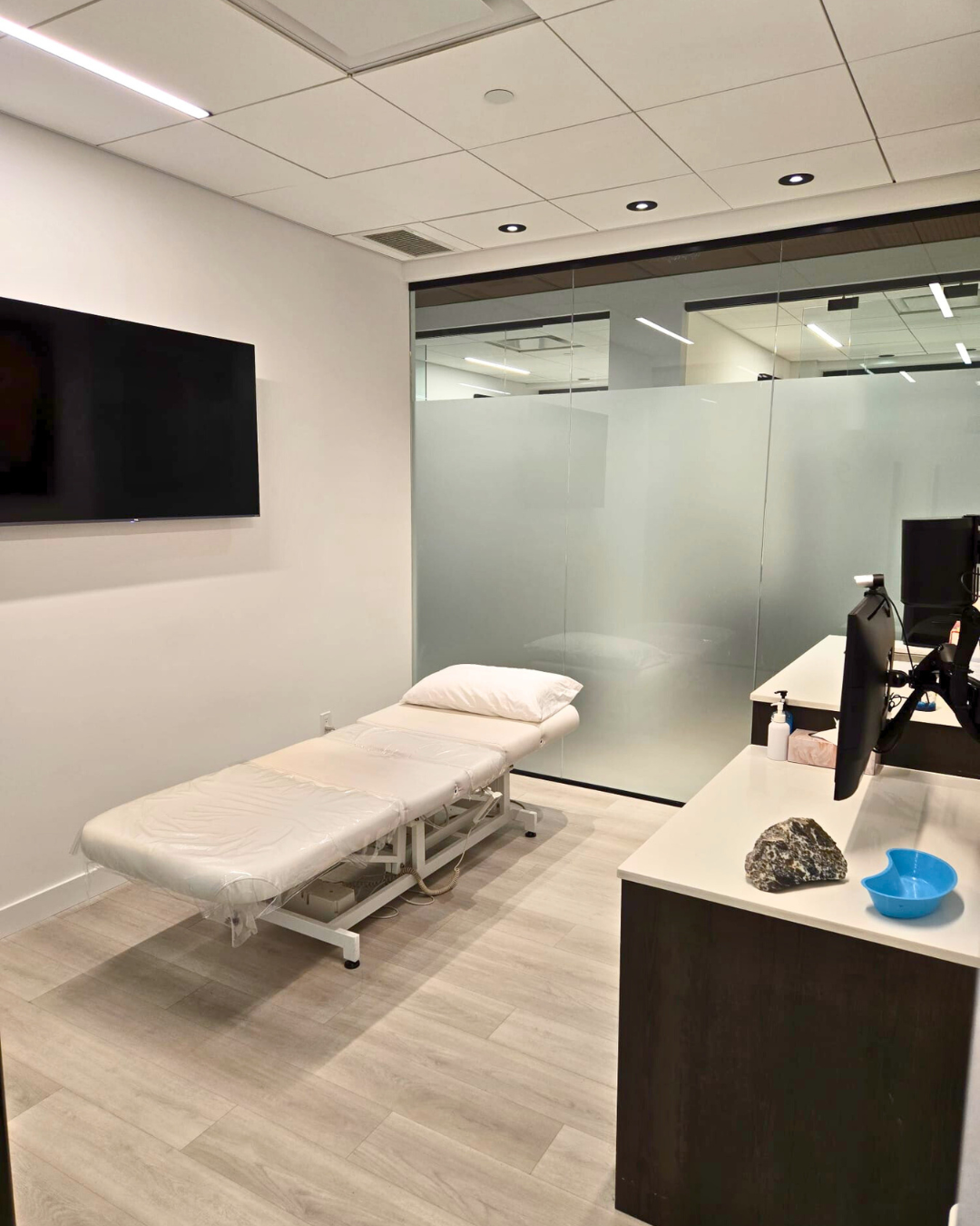 An empty medical examination room with a white examination bed, a pillow, a large flat-screen TV on the wall, and a desk with computer monitors, hand sanitizer, and a rock and blue bowl.