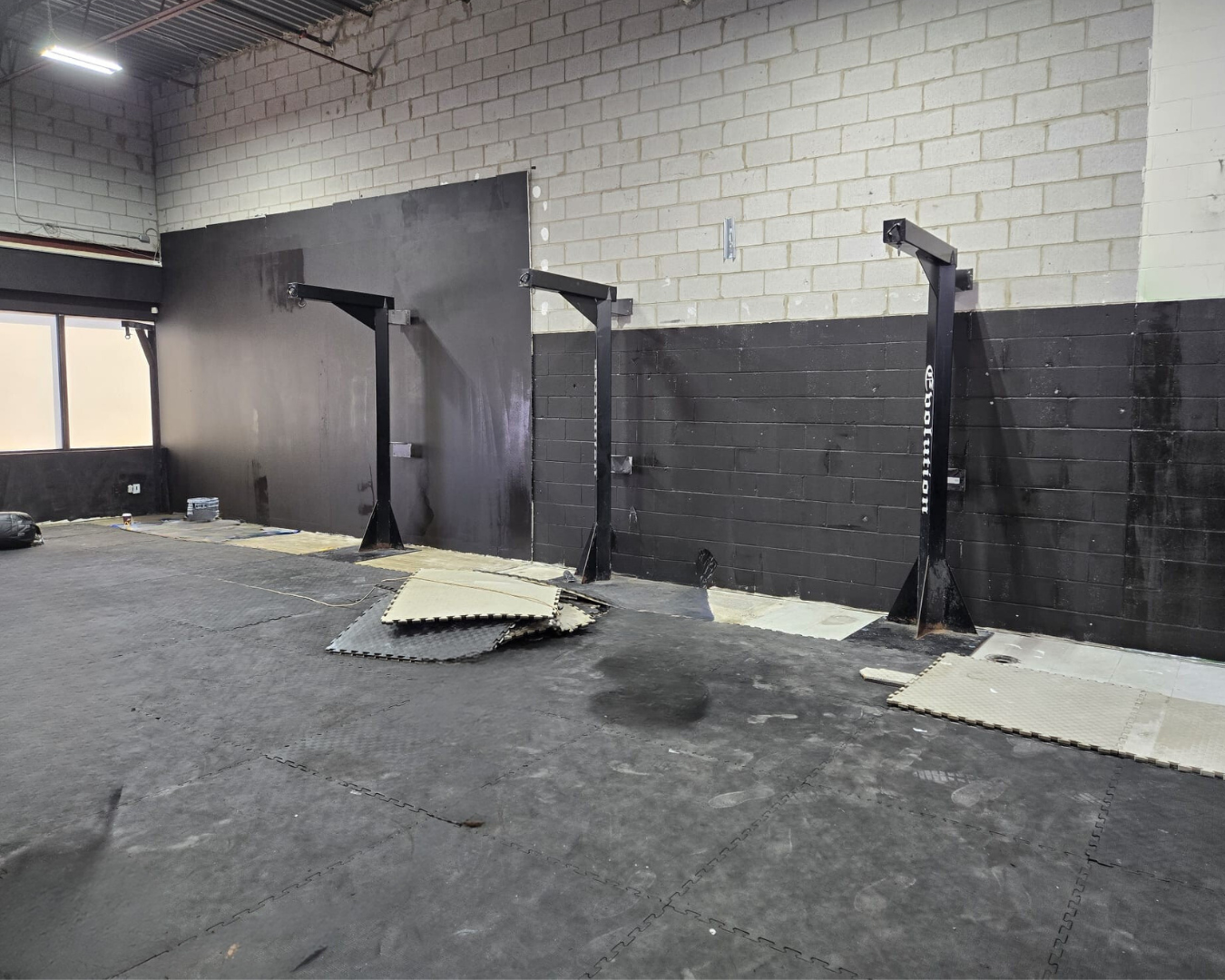 Indoor gym space with black wall, concrete floor with puzzle mats, and three black metal squat racks, some mats on the floor near the wall.