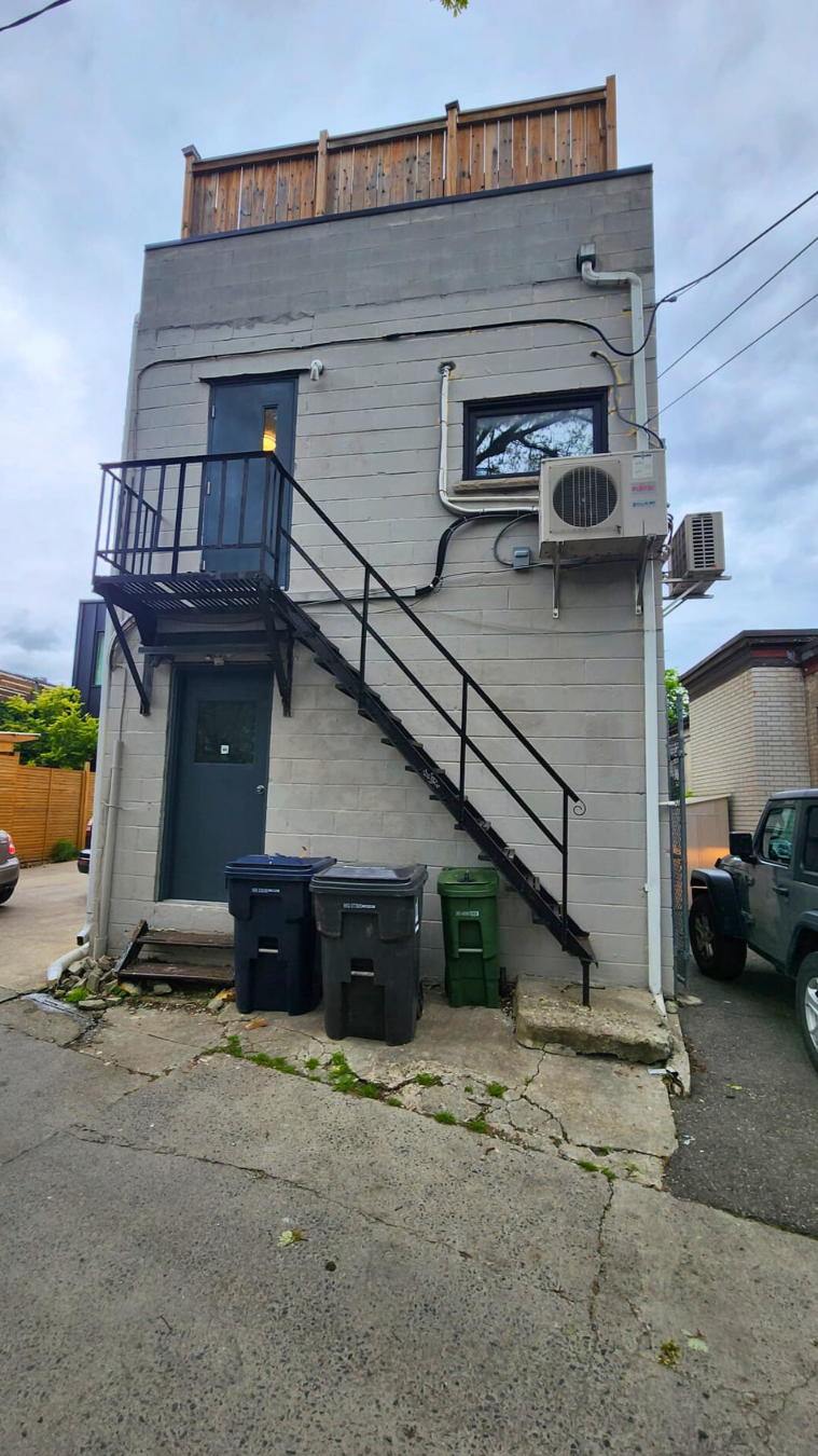 Back of a three-story building with a black metal exterior staircase, three trash cans in black, gray, and green at the base, a window and door on the ground level, two air conditioning units on the side wall, and a small balcony on the second floor. The building has a wooden fence on top and is situated on a cracked paved surface with some grass growing between the cracks.