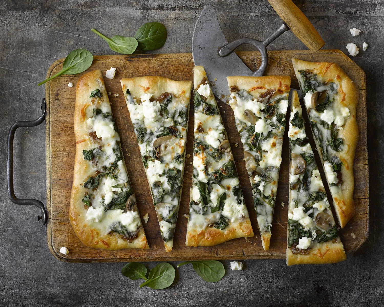 Slices of spinach and mushroom pizza on a wooden cutting board, with a pizza cutter and basil leaves nearby.
