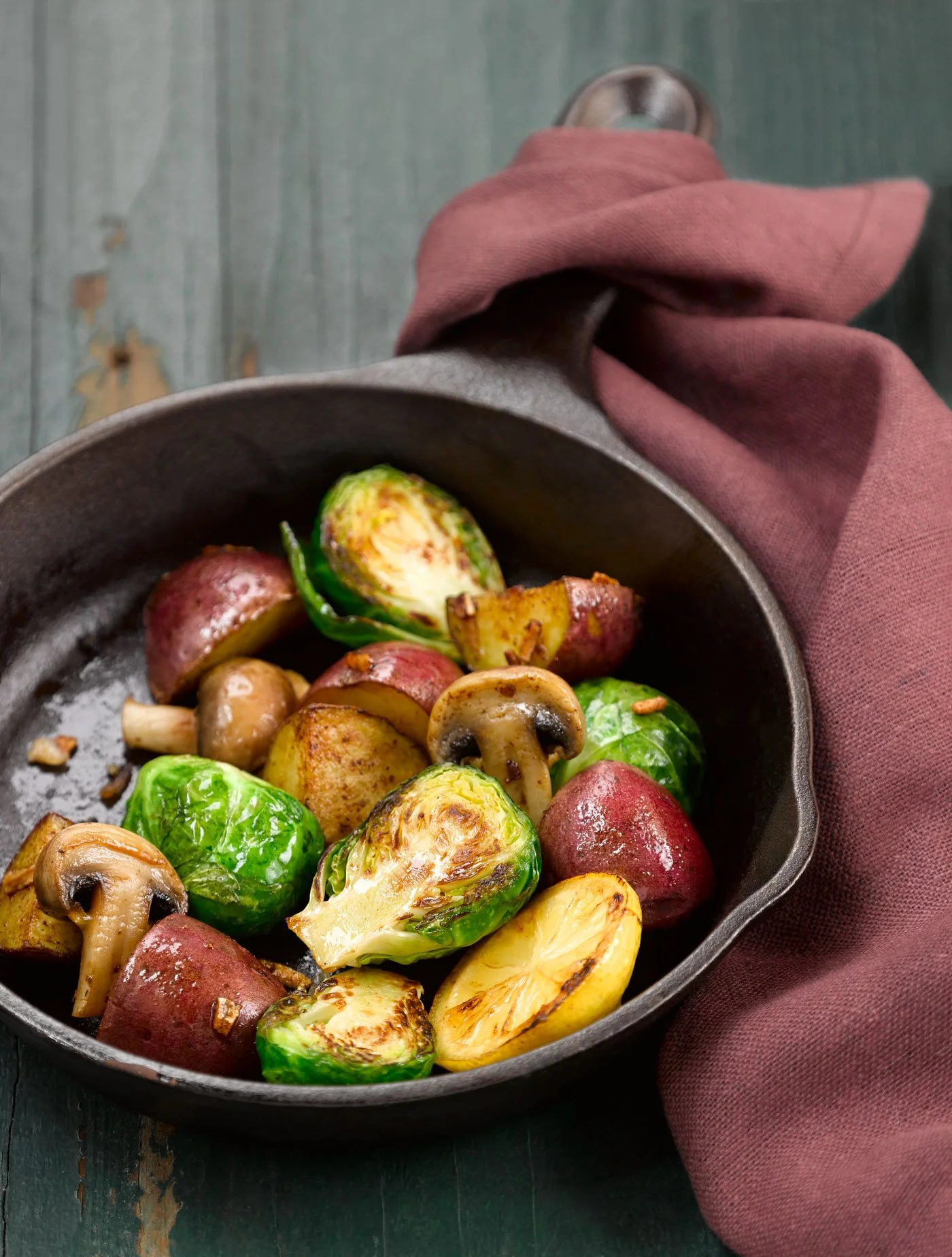 Cast iron skillet with roasted Brussels sprouts, red potatoes, yellow potatoes, and mushrooms, on a dark wooden surface with a maroon cloth.