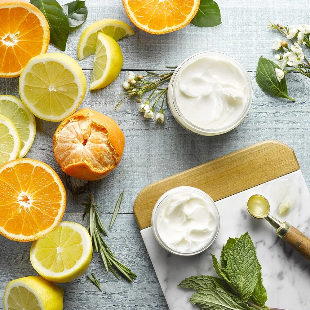 A flat lay of fresh citrus fruits, including oranges and lemons, along with jars of cream, sprigs of herbs, and a citrus juicer on a light wooden surface.