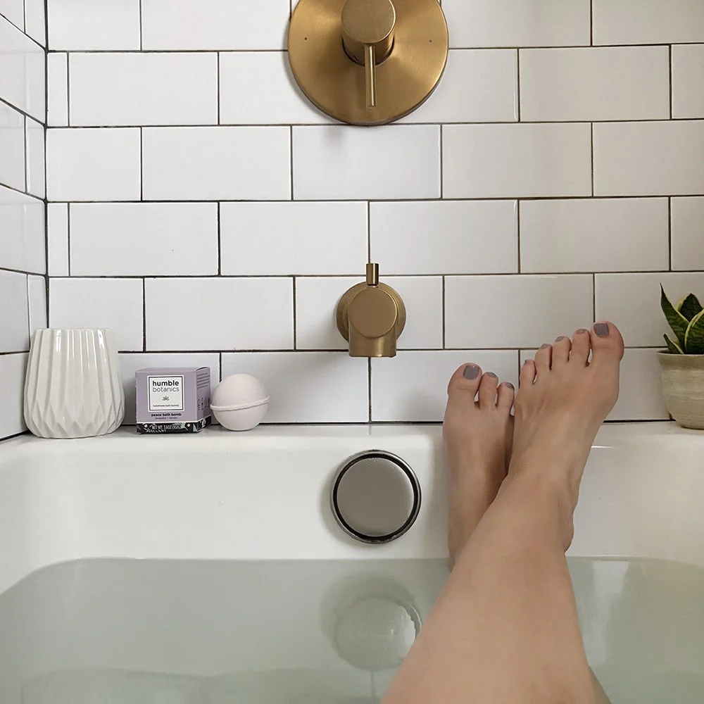 A person's legs resting on the edge of a bathtub filled with water, with various bath products and a small plant placed on the tub's edge. The tub's faucet and drain are visible against a white tiled wall.