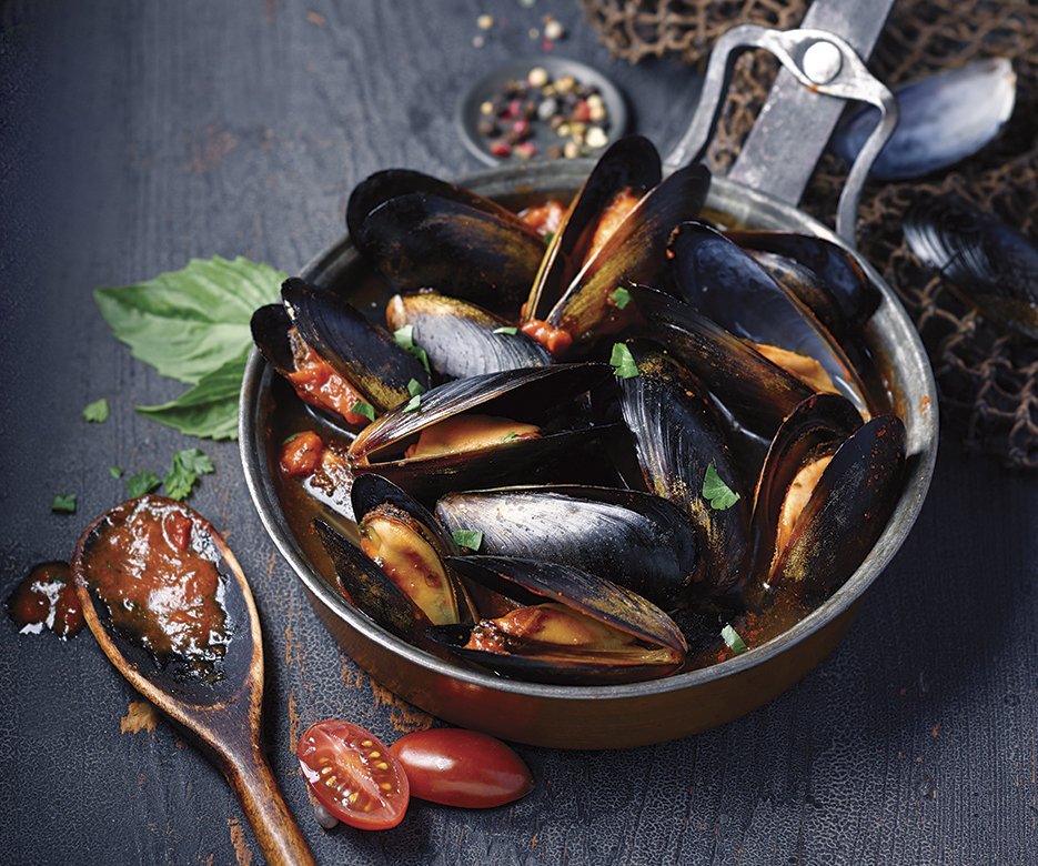 A pot of cooked mussels in tomato sauce garnished with chopped herbs, with cherry tomatoes and a basil leaf on the side, set on a dark surface.