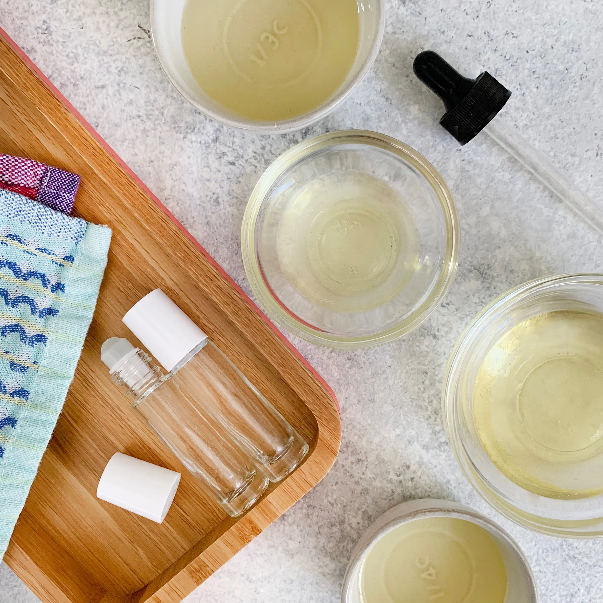 Several small glass bottles with white caps, a plastic spray bottle, a towel, and multiple containers with yellow liquid on a gray surface, with a bamboo tray nearby.