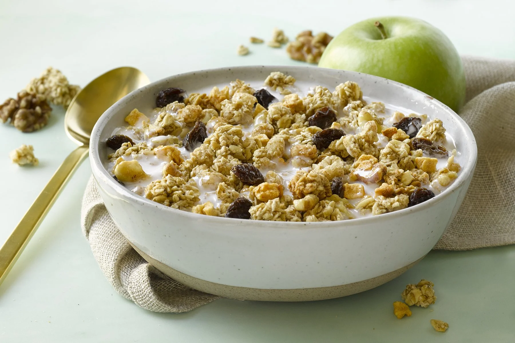 A bowl of granola with dried fruit and milk, next to a green apple, on a light green surface.