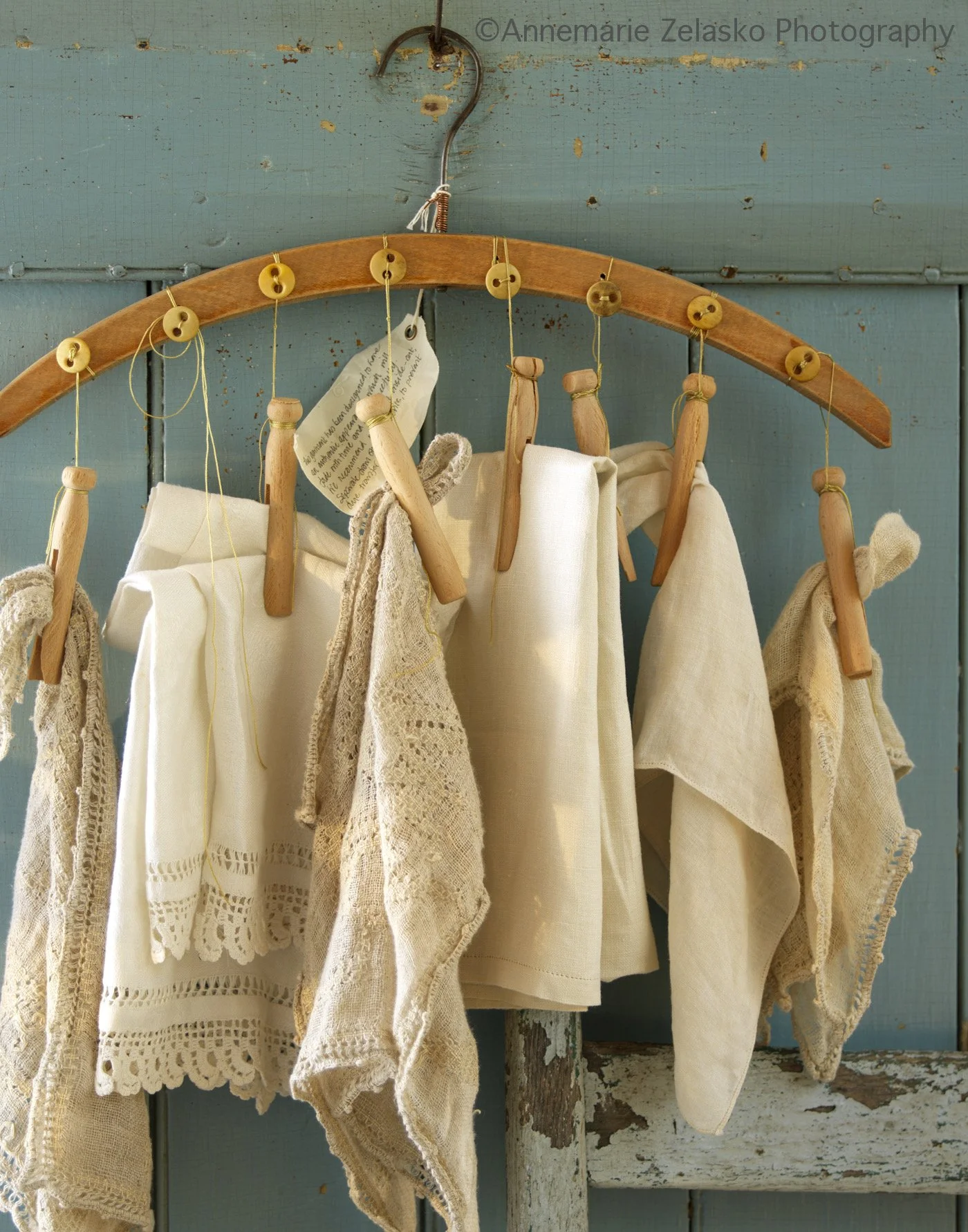 A wooden hanger holding vintage cream-colored fabric pieces and lace cloths against a weathered teal wall.