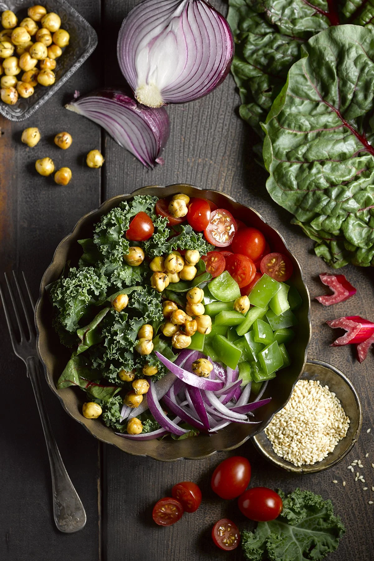 A bowl of fresh chopped vegetables, including cherry tomatoes, sliced bell peppers, sliced red onions, kale, and chickpeas, on a wooden surface with additional vegetables and chickpeas scattered around.