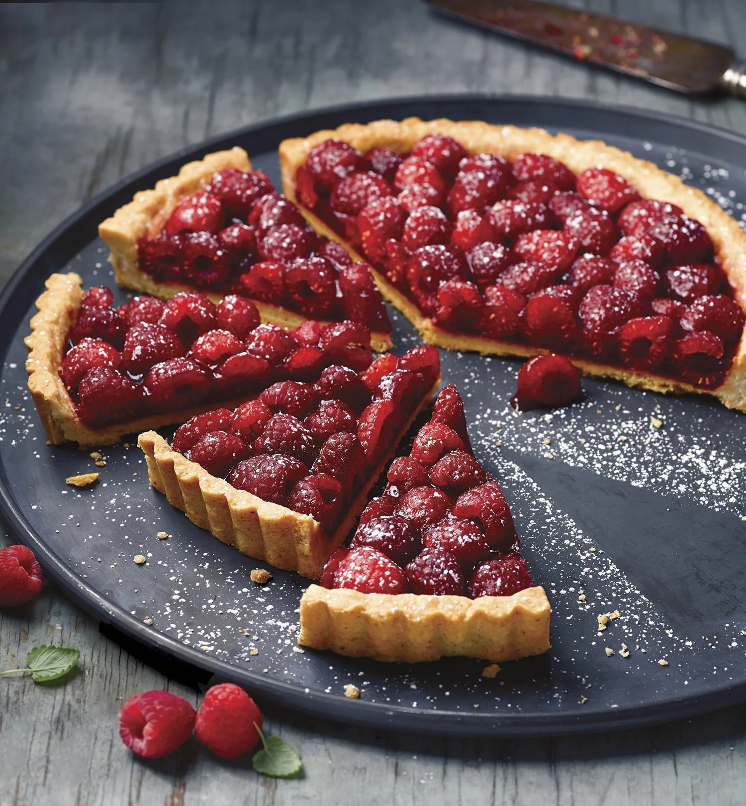 A raspberry tart with a golden crust and fresh raspberries, dusted with powdered sugar on a dark plate