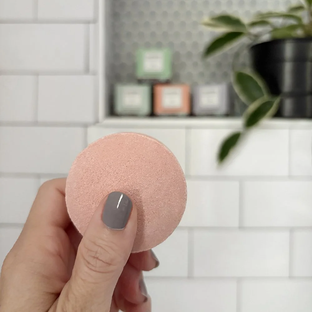 Hand holding a pink makeup sponge in front of a white tiled wall, with a potted plant and blurred boxes in the background.