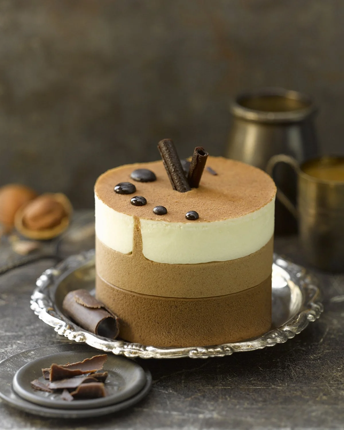 A layered chocolate and vanilla mousse cake with chocolate shavings and coffee beans on top, placed on a silver tray with additional chocolate pieces nearby.