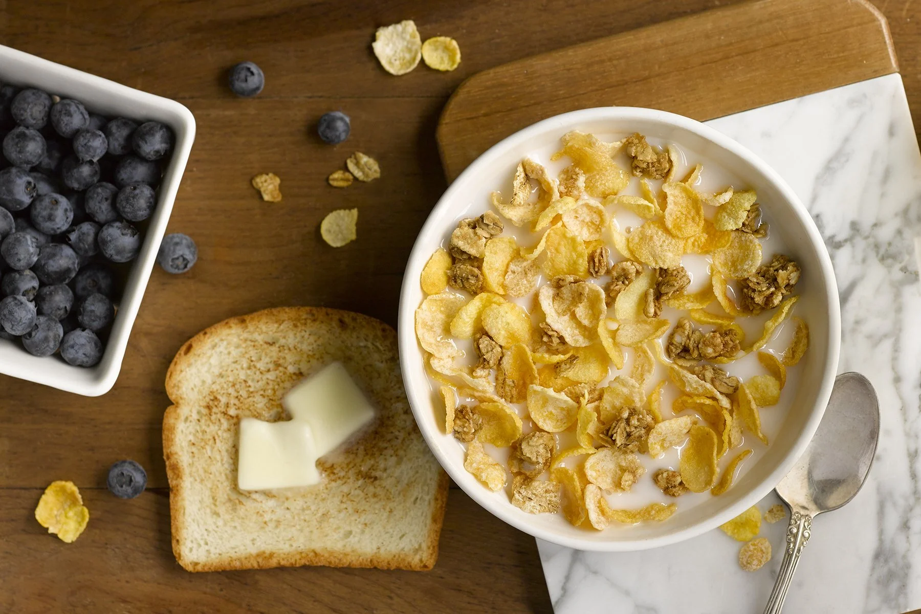 A bowl of cereal with milk topped with cornflakes and walnuts, a slice of toasted bread with butter, a dish of blueberries, and a silver spoon on a wooden table.