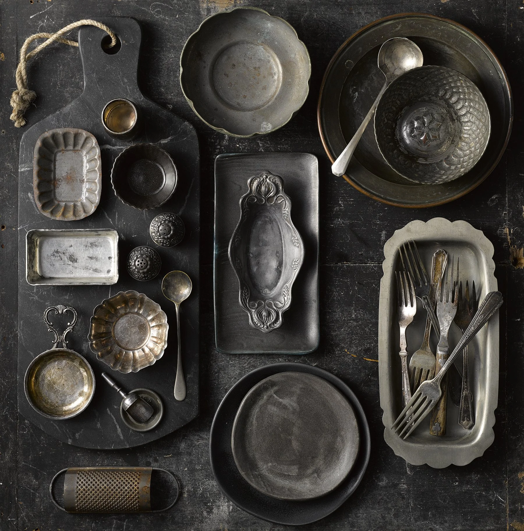 Collection of vintage metal and ceramic dishes, bowls, trays, and utensils arranged on a dark surface.