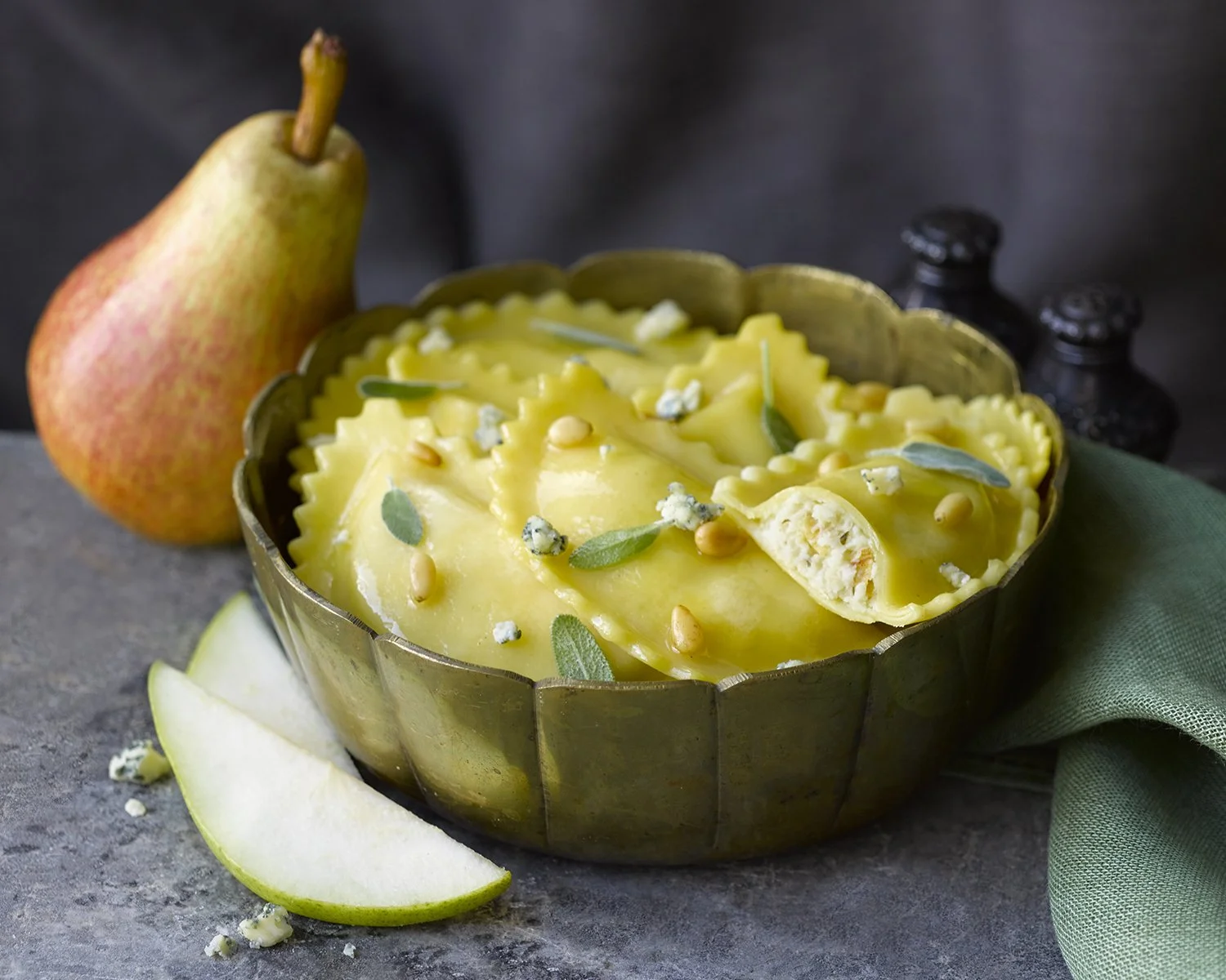 Slices of pear and ravioli topped with yellow sauce, garnished with blue cheese crumbles, sage leaves, and pine nuts in a brass bowl, with a pear, green cloth, and salt and pepper shakers in the background.