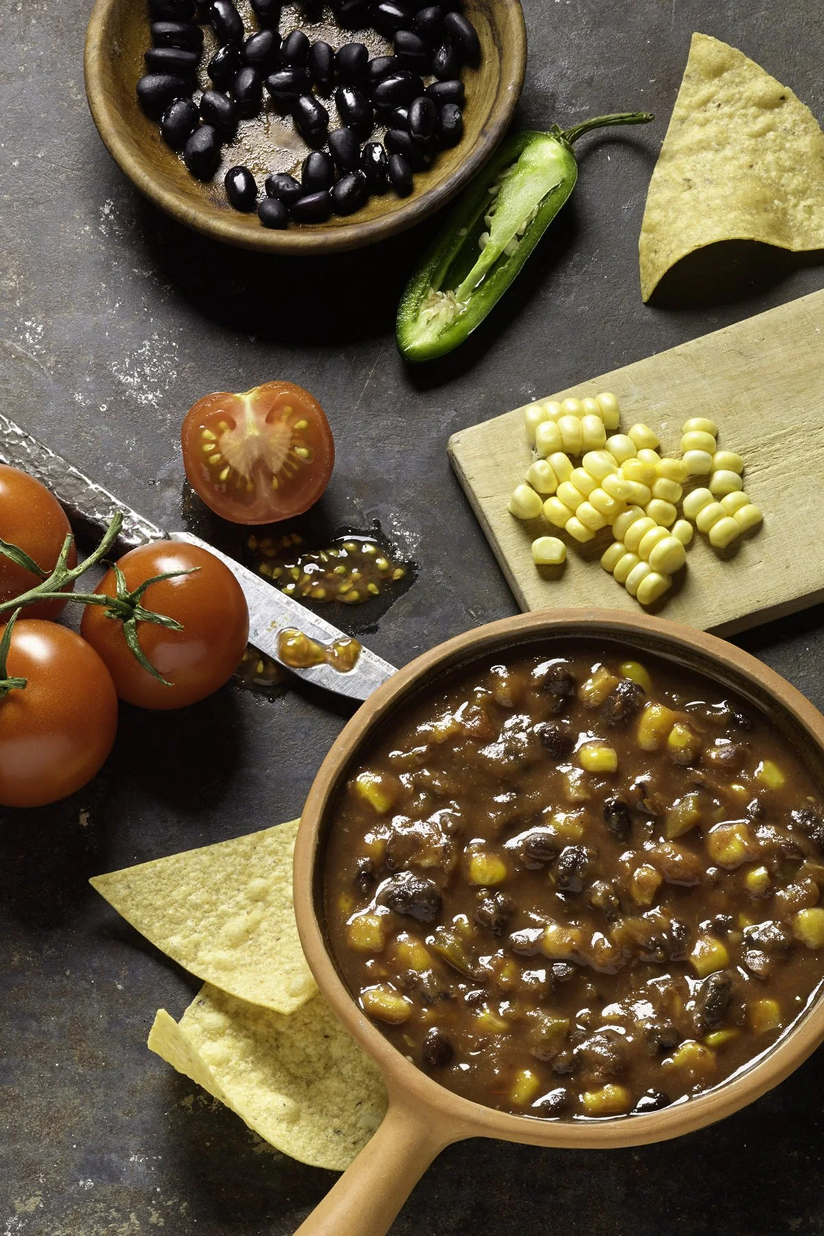 A bowl of chili with black beans and corn next to fresh tomatoes, jalapeno pepper, tortilla chips, and a wooden bowl filled with black beans on a dark surface.