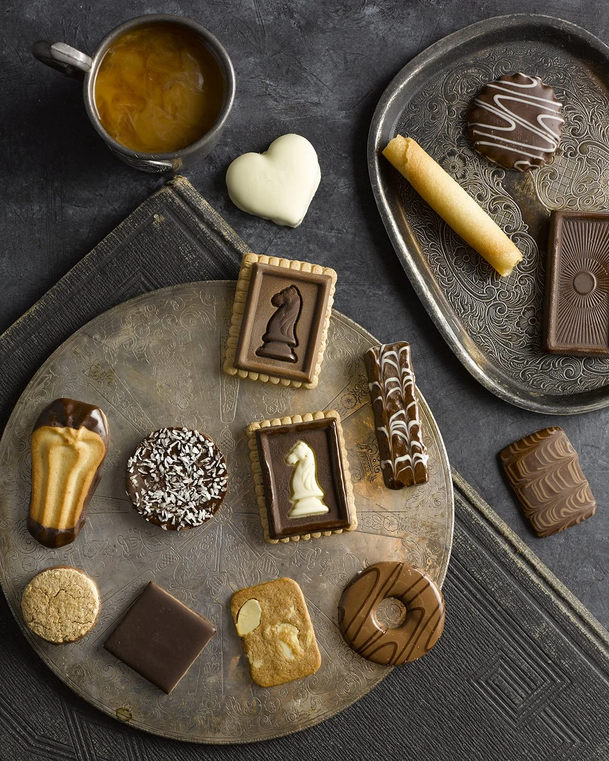 Assorted chocolates and cookies on a gold decorative plate, with a cup of tea or coffee and a white heart-shaped confection nearby.