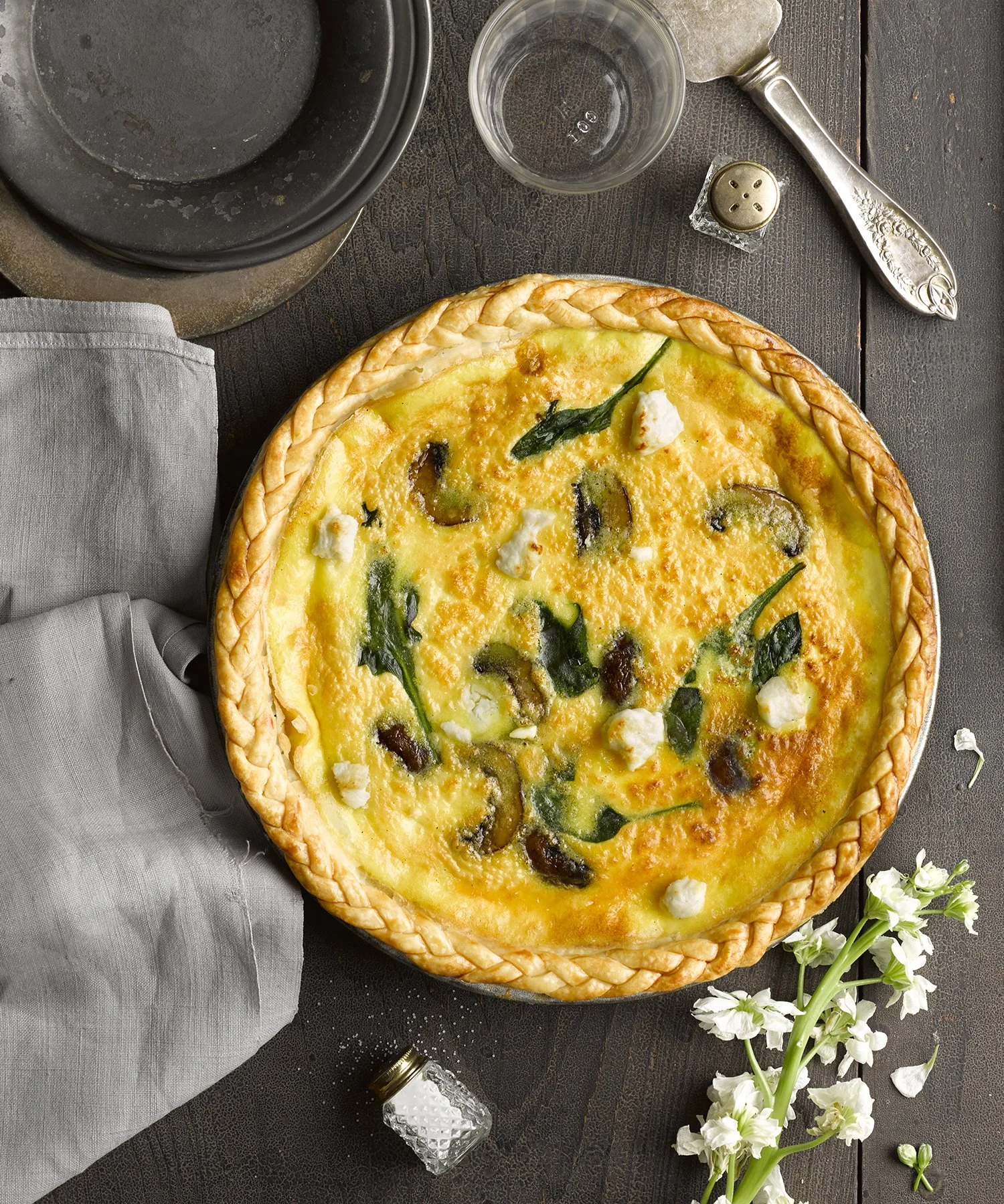 A cheese and vegetable pizza with a braided crust on a dark wooden table, accompanied by a glass of water, a pepper shaker, a napkin, a small dish with salt, and a flower.