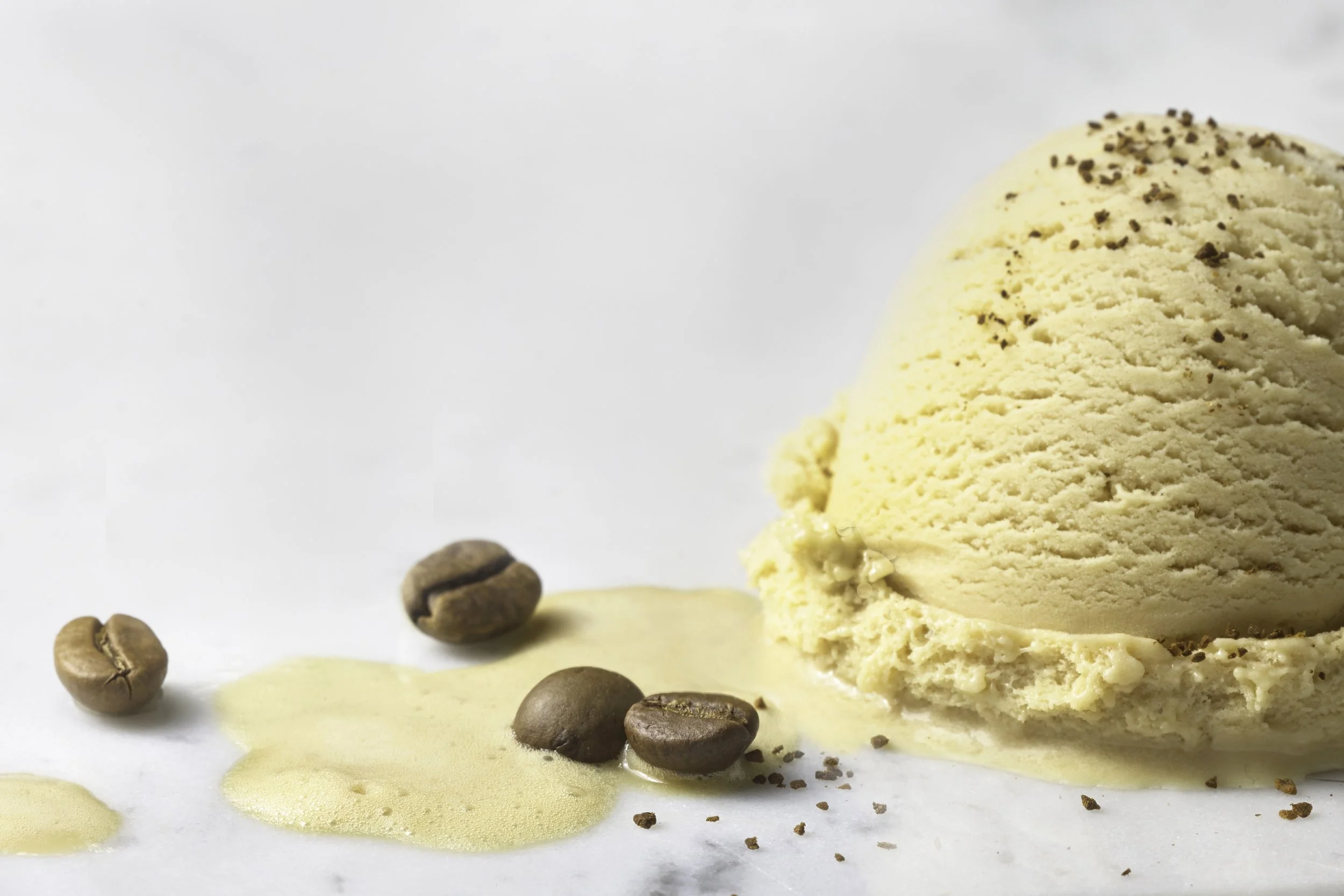A scoop of vanilla ice cream with coffee beans and spilled coffee on a white surface.