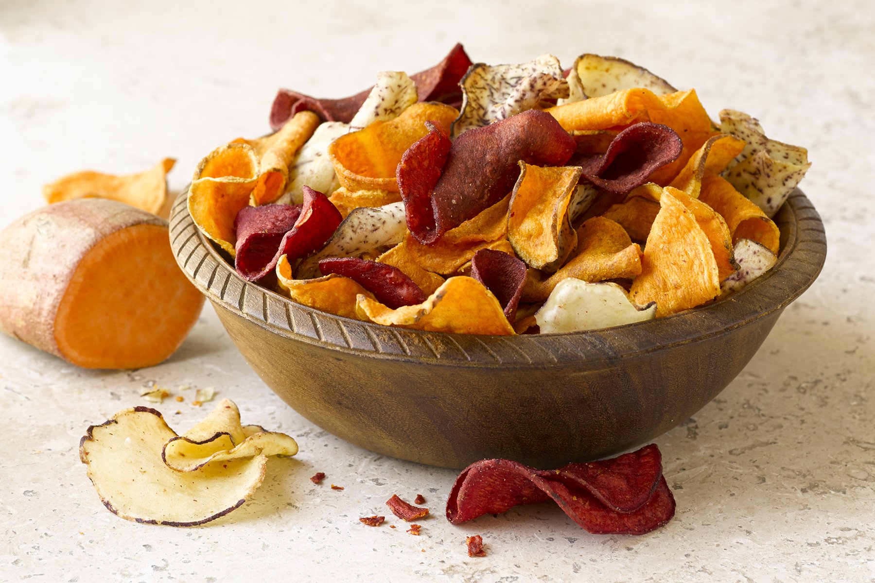 A wooden bowl filled with colorful vegetable chips including sweet potato, beet, and parsnip chips, with some chips and a sliced sweet potato on a light-colored surface nearby.