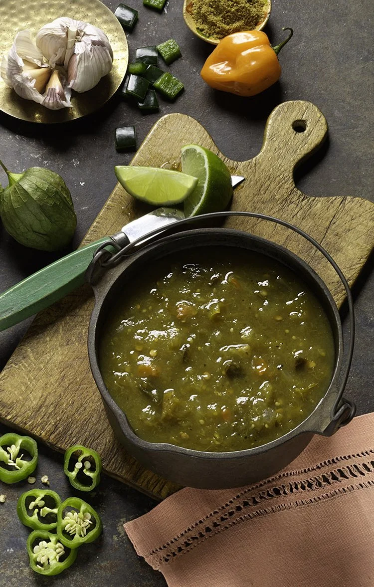 A black pot of hot vegetable soup on a wooden cutting board surrounded by lime wedges, chopped green peppers, garlic, a green tomatillo, a small bowl of chili powder, and a yellow and orange bell pepper.