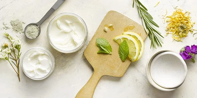 Lemon slices, mint, ginger, and rosemary on a cutting board, with yogurt and herbs in bowls on a white surface.