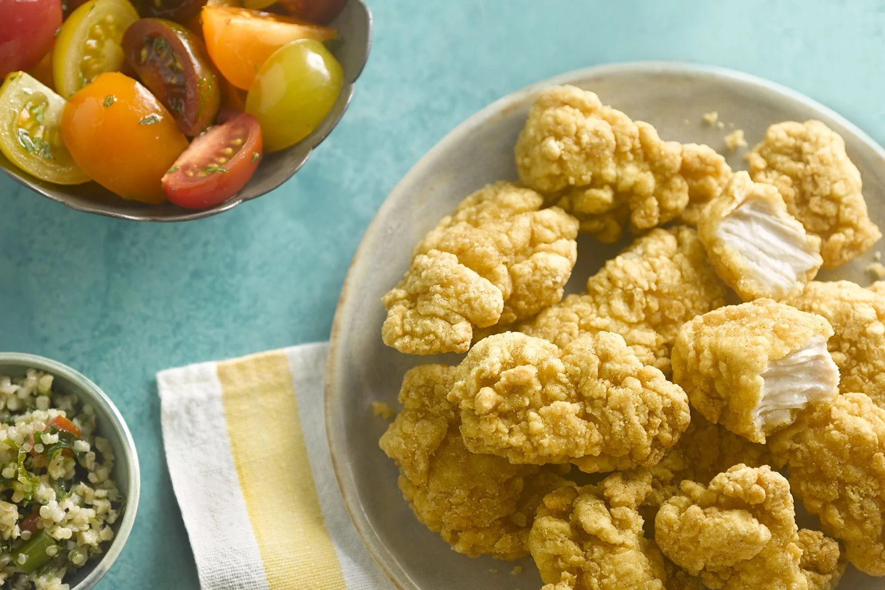 Plate of fried chicken tenders with a side of cherry tomato salad and a small bowl of grain salad on a turquoise table.