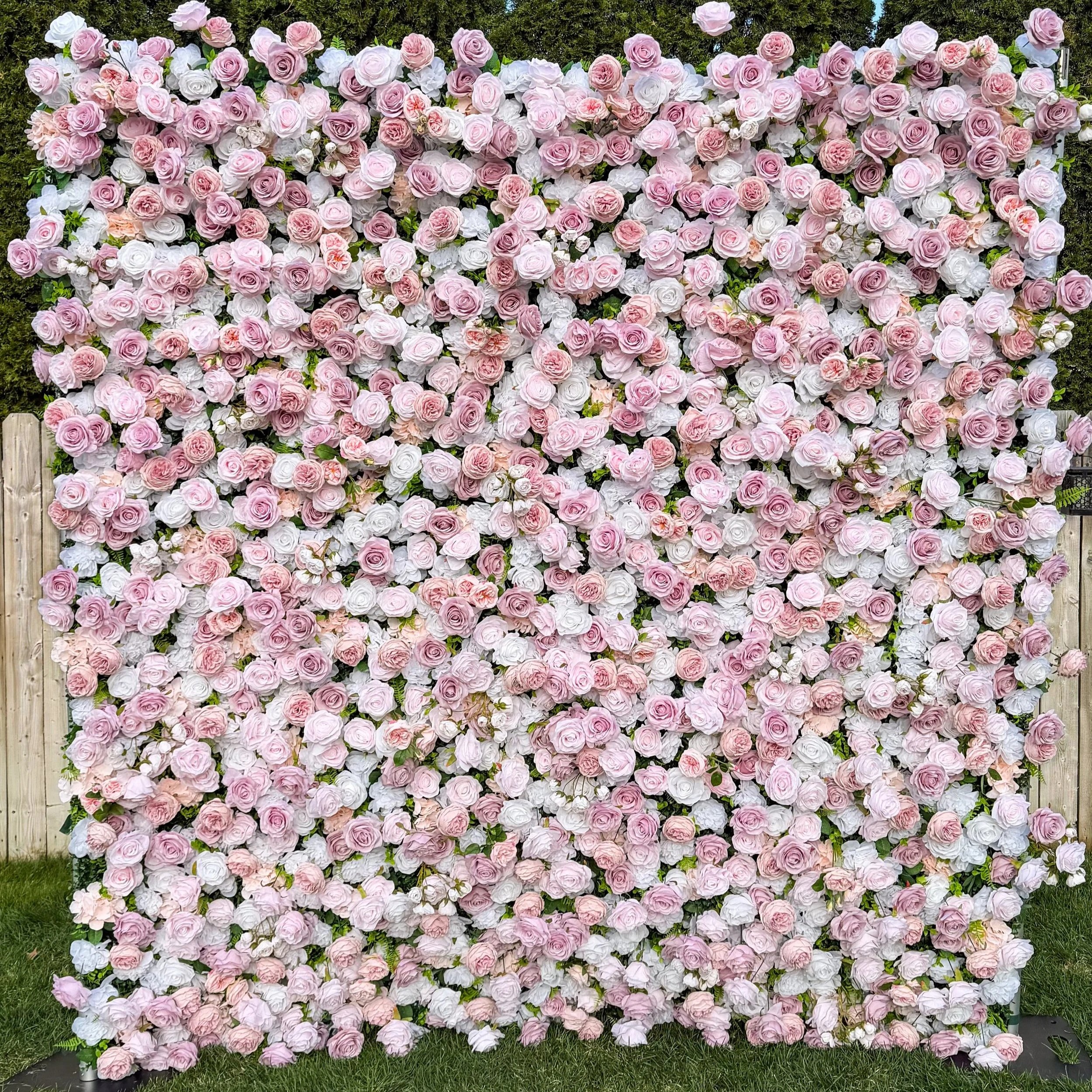 A large display of pink and white roses arranged on a floral wall with a wooden fence at the bottom and greenery in the background.