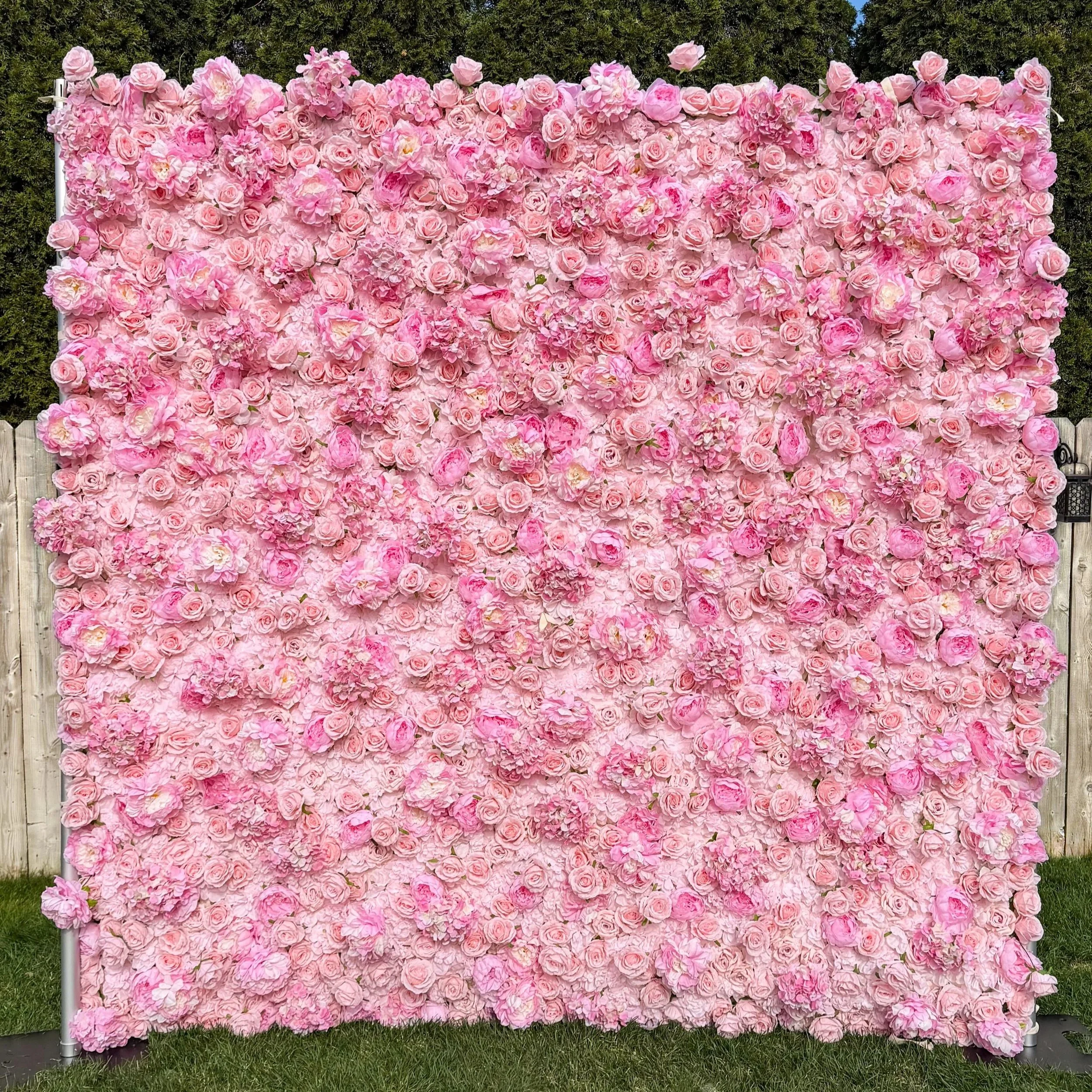 A large floral wall made of pink roses and peonies arranged outdoors on a metal frame, with a background of trees and blue sky.