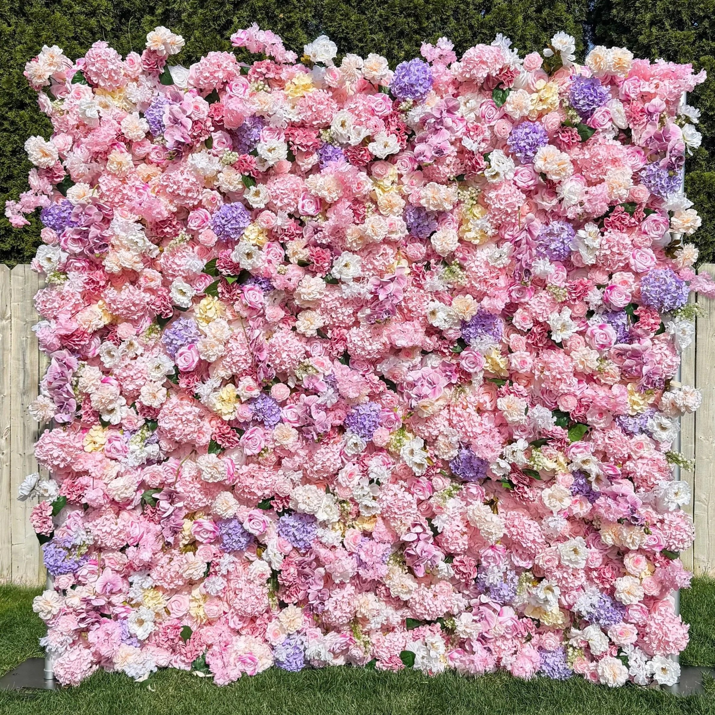 A wall covered with pink, purple, and white hydrangea and rose flowers, set outdoors on a grassy area with a wooden fence at the bottom.