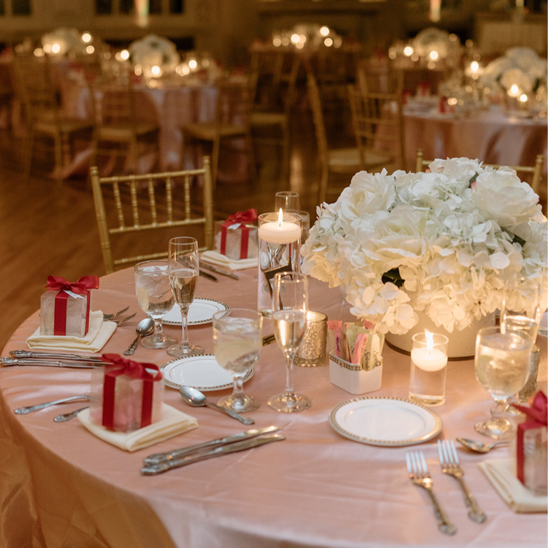 Round banquet table with a pink tablecloth, decorated with a large white floral centerpiece, candles, wine glasses, plates, silverware, and small gift boxes with red ribbons. Background shows other tables and chairs in an elegant event hall.