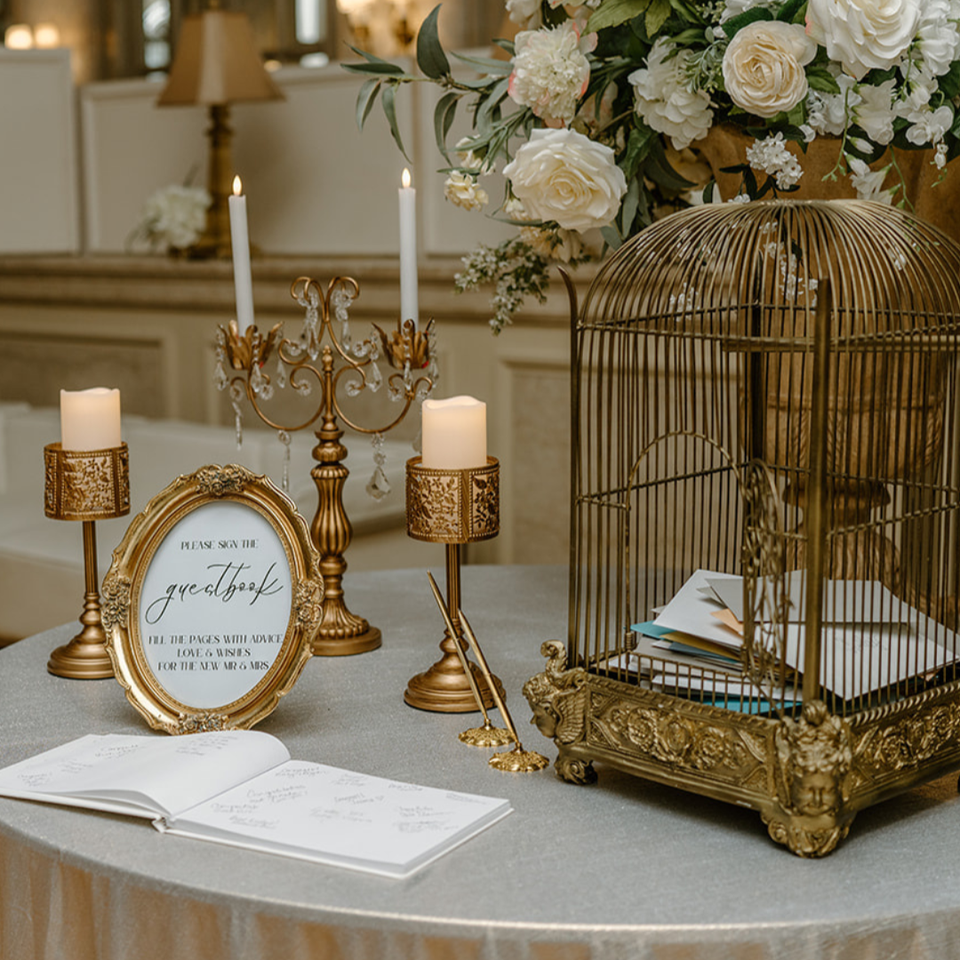 A wedding guestbook table with gold accents, featuring a guestbook, pen, a gold birdcage filled with paper, a sign asking guests to sign the guestbook, candles, and a floral arrangement with white roses and greenery.