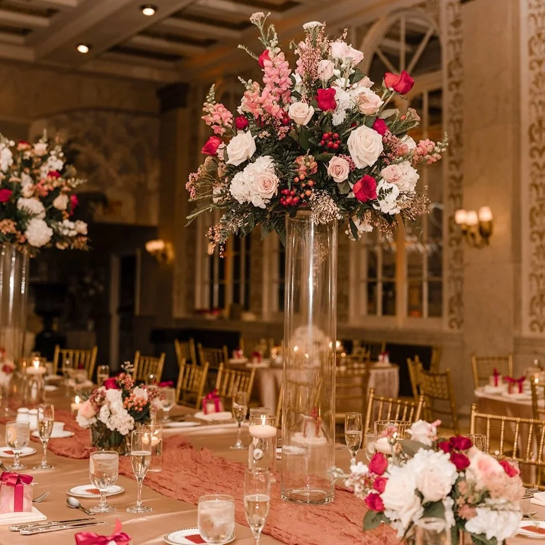 A banquet hall decorated with tall floral centerpieces featuring pink, white, and red flowers on tables set with plates, glasses, and wrapped gifts.