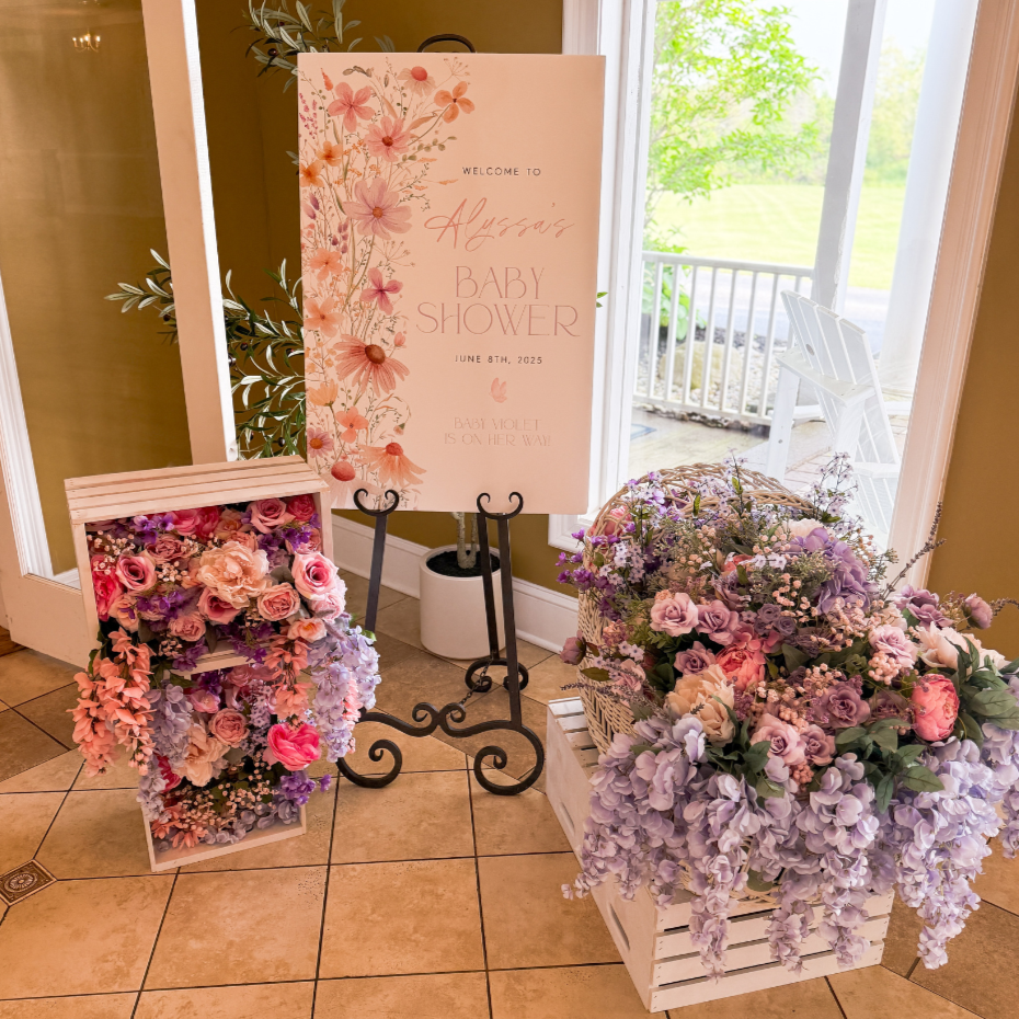 A floral display with pink and purple flowers, a welcome sign for Alberta's baby shower, and a basket of flowers near an open door with a view of a porch and green outdoors.