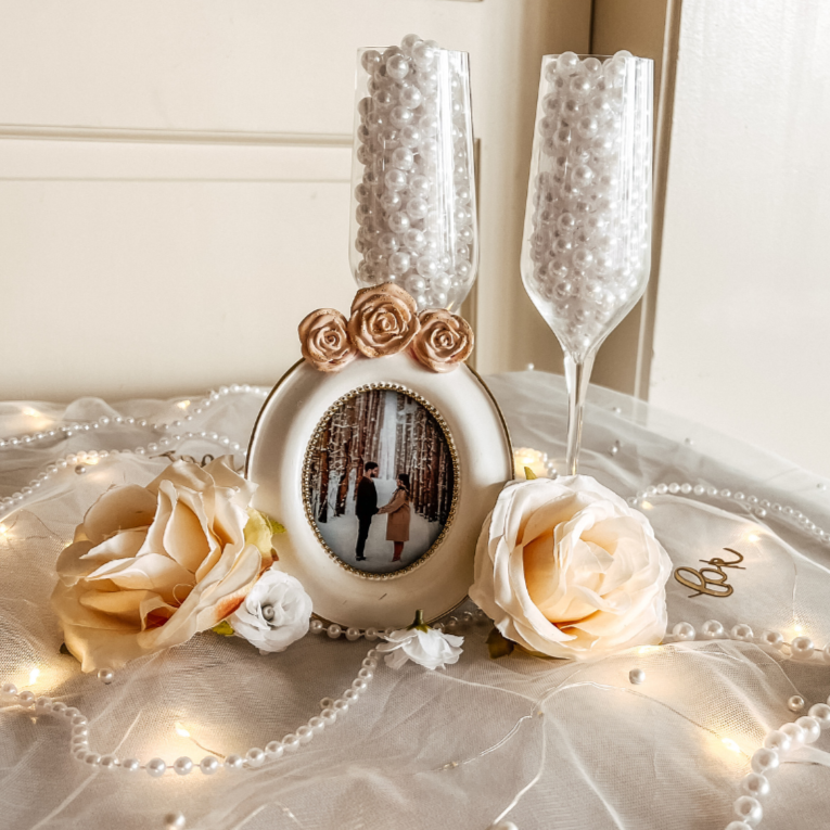 Decorative wedding display with a picture frame, artificial cream-colored roses, pearl strings, and two champagne glasses filled with white beads, on a white cloth.