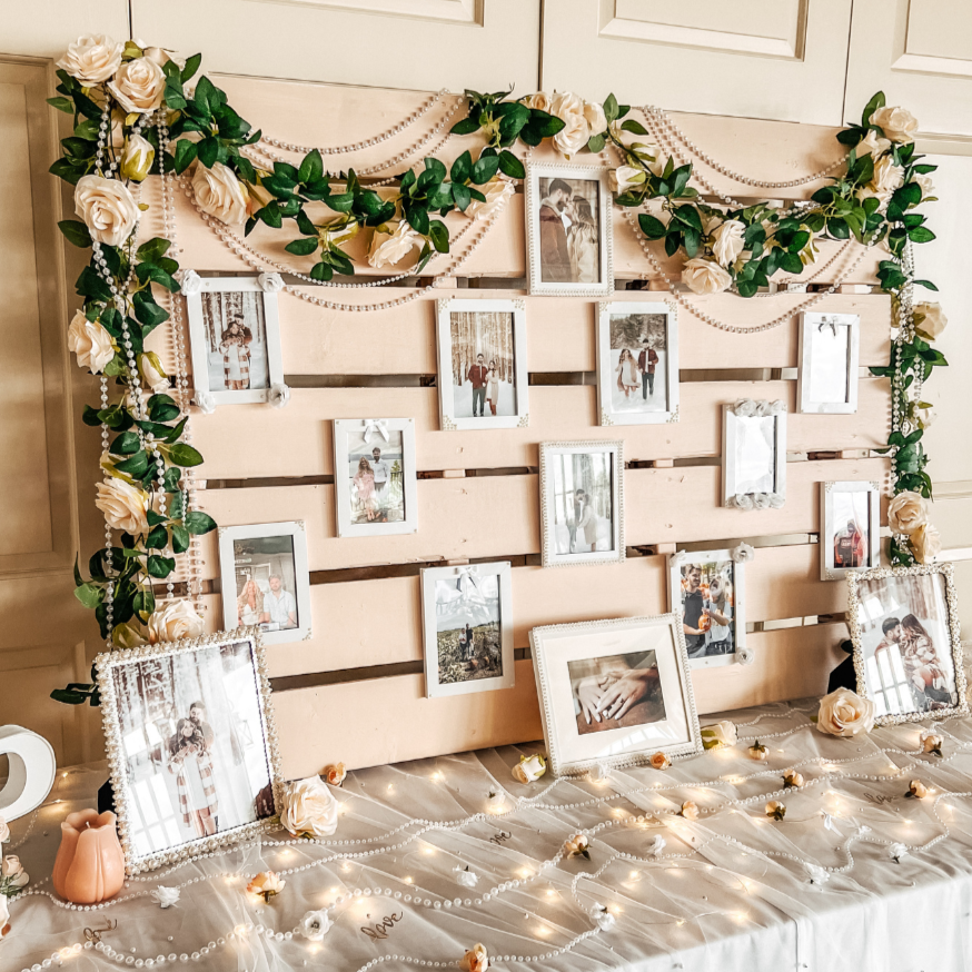 Decorative photo display with framed pictures of couples and individuals, adorned with faux roses, green leaves, pearl garlands, and string lights on a wooden backdrop, possibly for a wedding or special event.