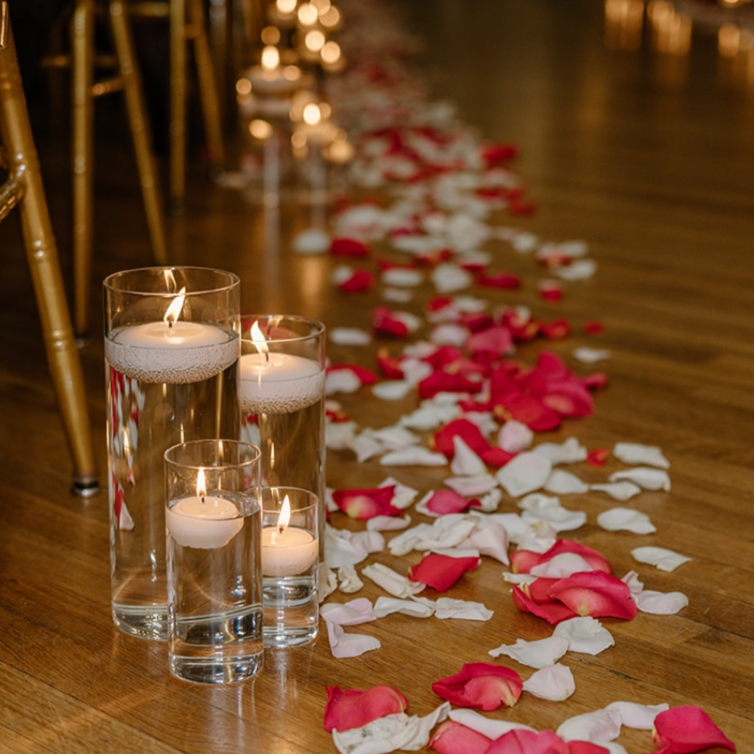Four tall glass candles with floating flames and one small candle on a wooden floor, surrounded by scattered pink and white rose petals, forming a path. Gold chairs are visible along the side in a dimly lit setting.