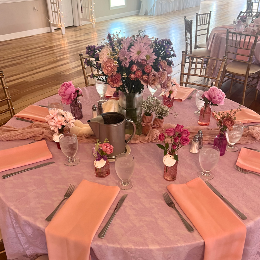 Round table decorated with a pink tablecloth, pink napkins, and pink and purple floral centerpieces, set for a formal event in a spacious hall with wooden floors and empty chairs in the background.