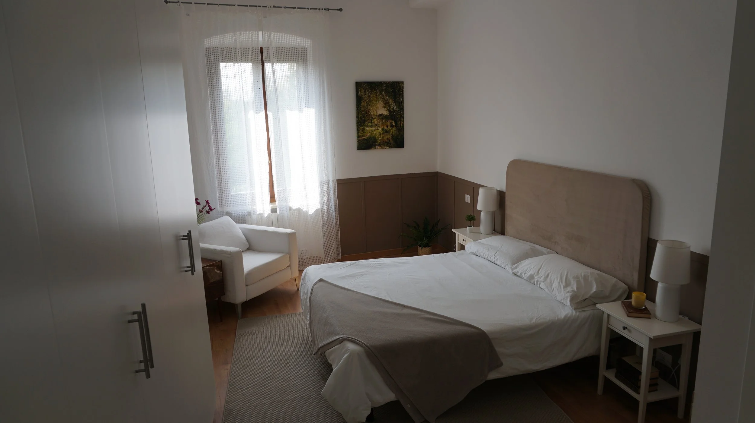 Camera da letto luminosa con letto matrimoniale, due lampade da notte, un quadro su una parete e una sedia bianca vicino alla finestra con tende leggere.