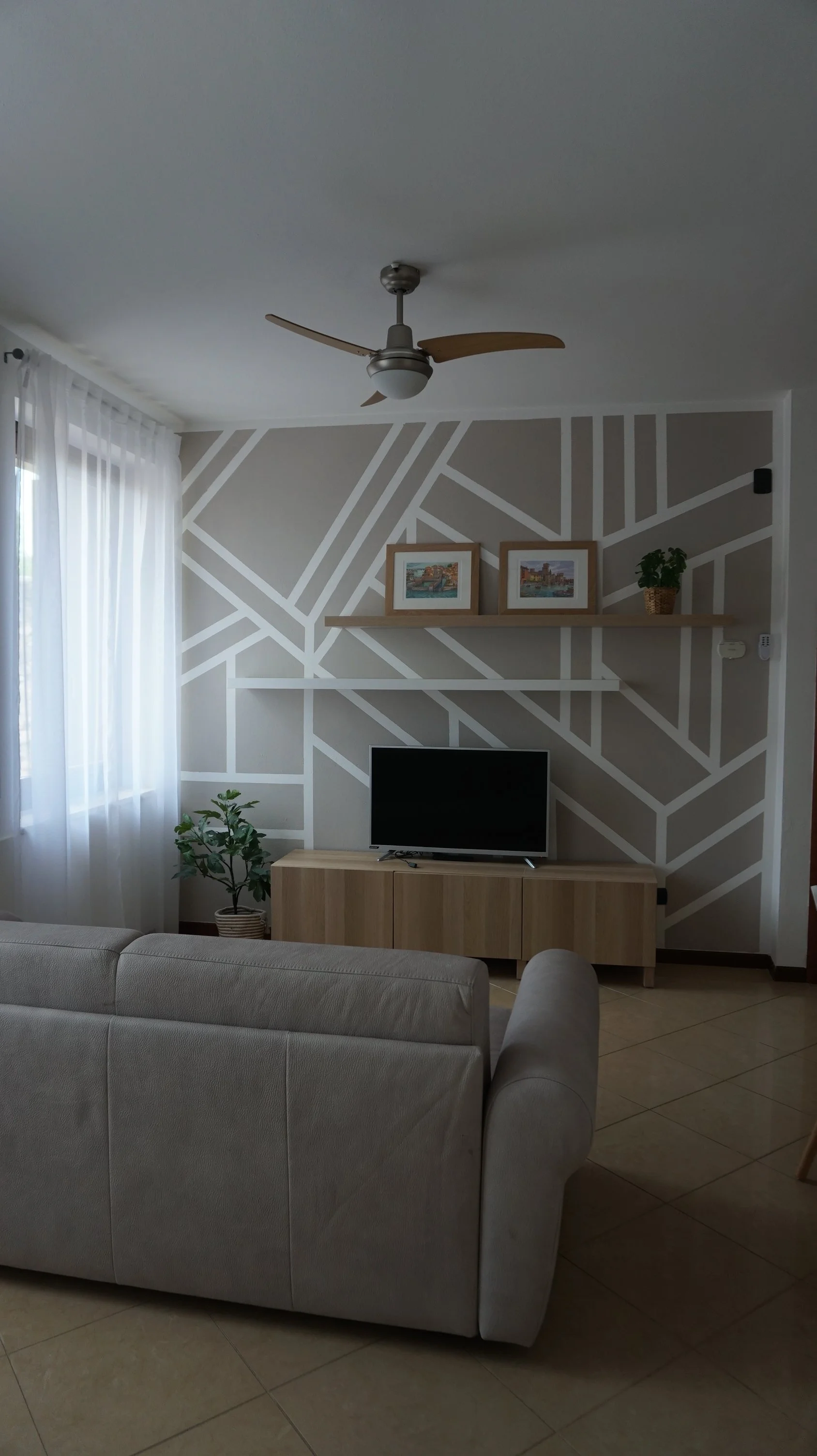 Interno di un soggiorno con pareti decorate con strisce bianche su sfondo beige, un televisore su un mobile in legno, una pianta in vaso e due quadri su una mensola sopra il televisore.
