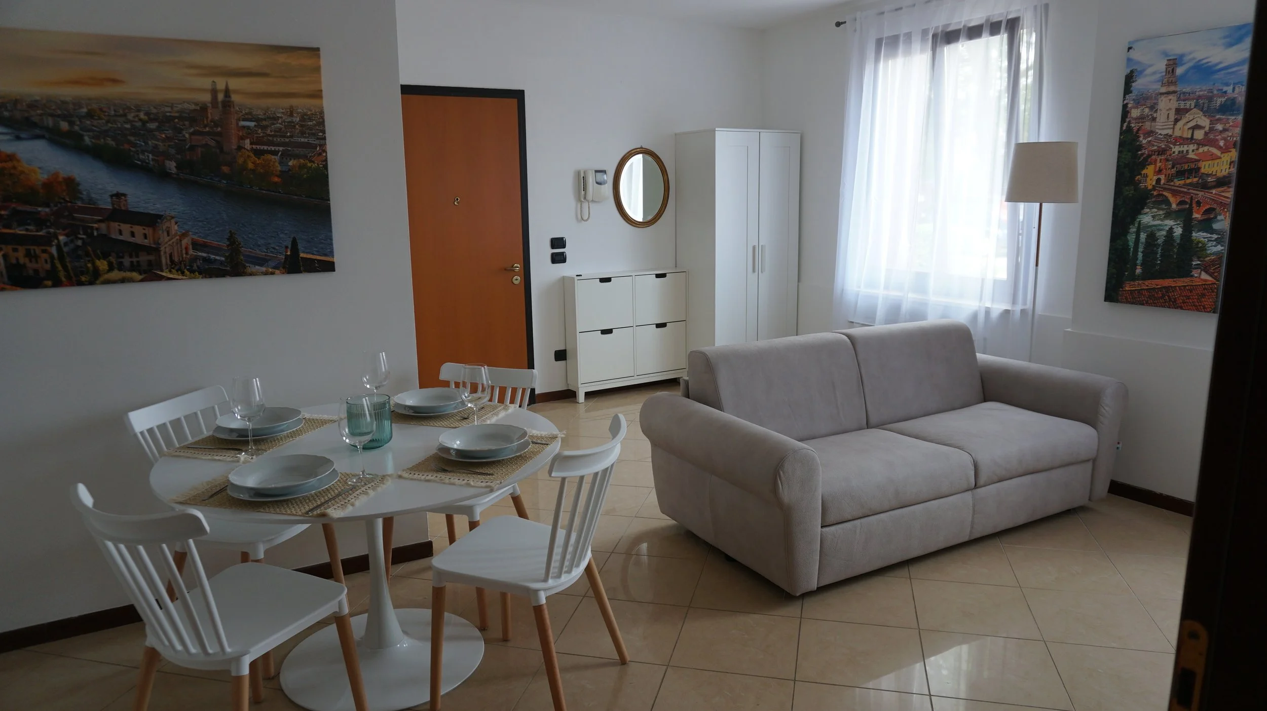 Soggiorno con cucina moderna, divano beige, tavolo da pranzo rotondo con sedie bianche, quadri verdi e blu alle pareti, finestra con tende leggere, porta in legno.