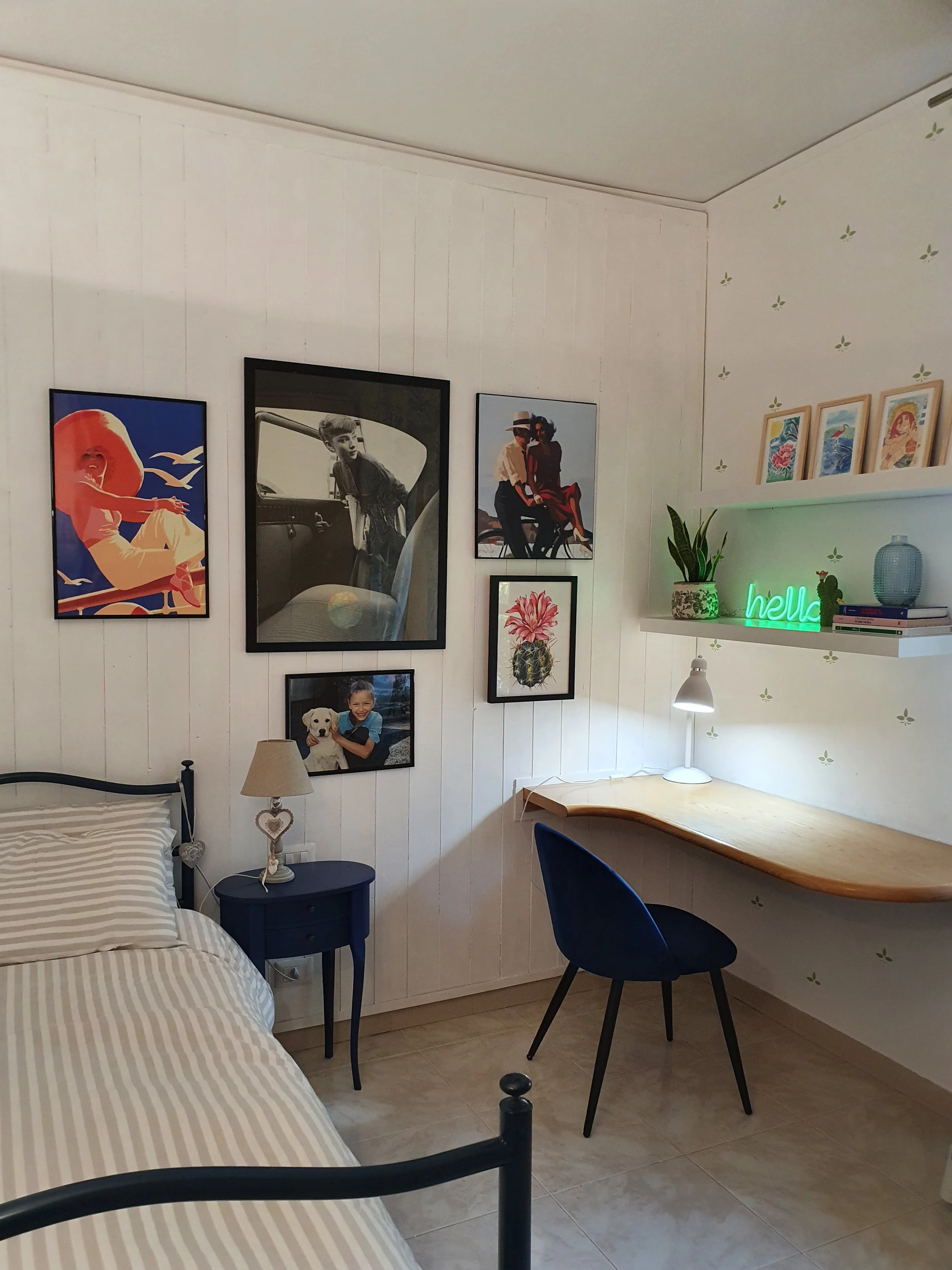 Stanza con pareti decorate con quadri, una scrivania con sedia, lampada da scrivania e mensola con decorazioni e libri.