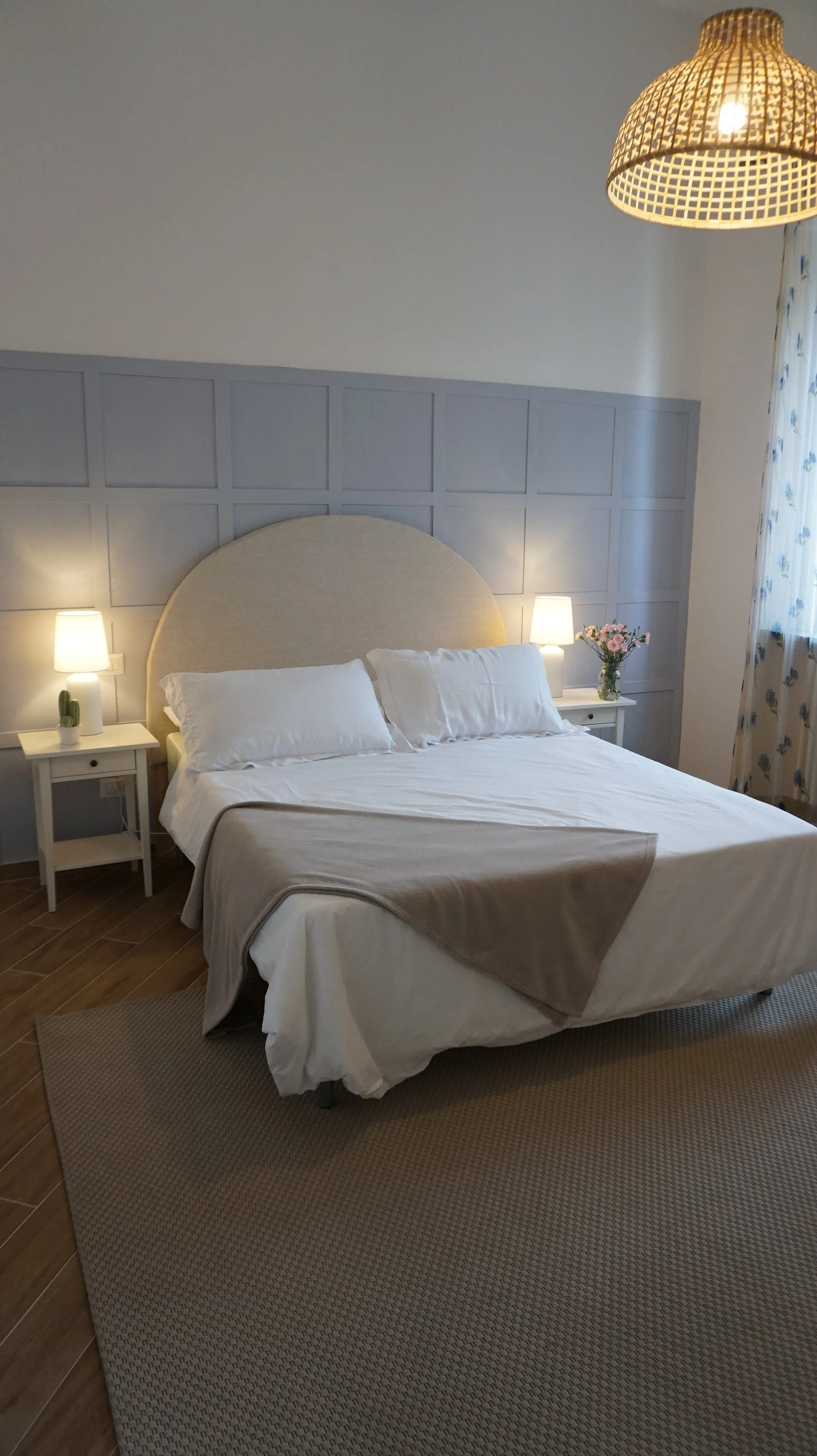 Letto matrimoniale in una stanza con luci laterali accese, candelella con cactus e vaso di fiori su comodino, pareti grigie, tappeto beige, tende con stampa floreale.