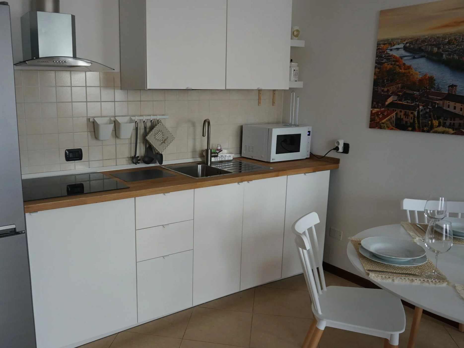 Cucina moderna con mobili bianchi, piano di lavoro in legno, fornello elettrico, lavello, microonde e un'illustrazione di una città sul muro.