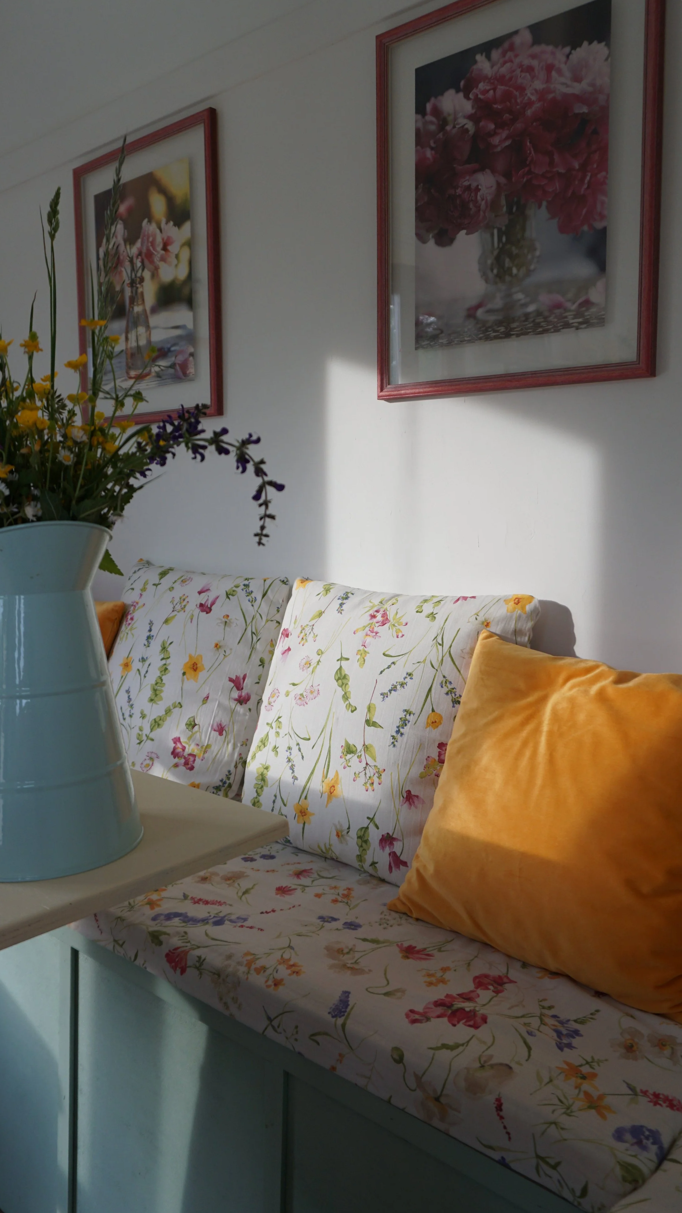 Un divano con cuscini con fodere floreali, un vaso con fiori, quadri appesi al muro con immagini di fiori, e una sorgente di luce naturale che crea ombre sulla parete.