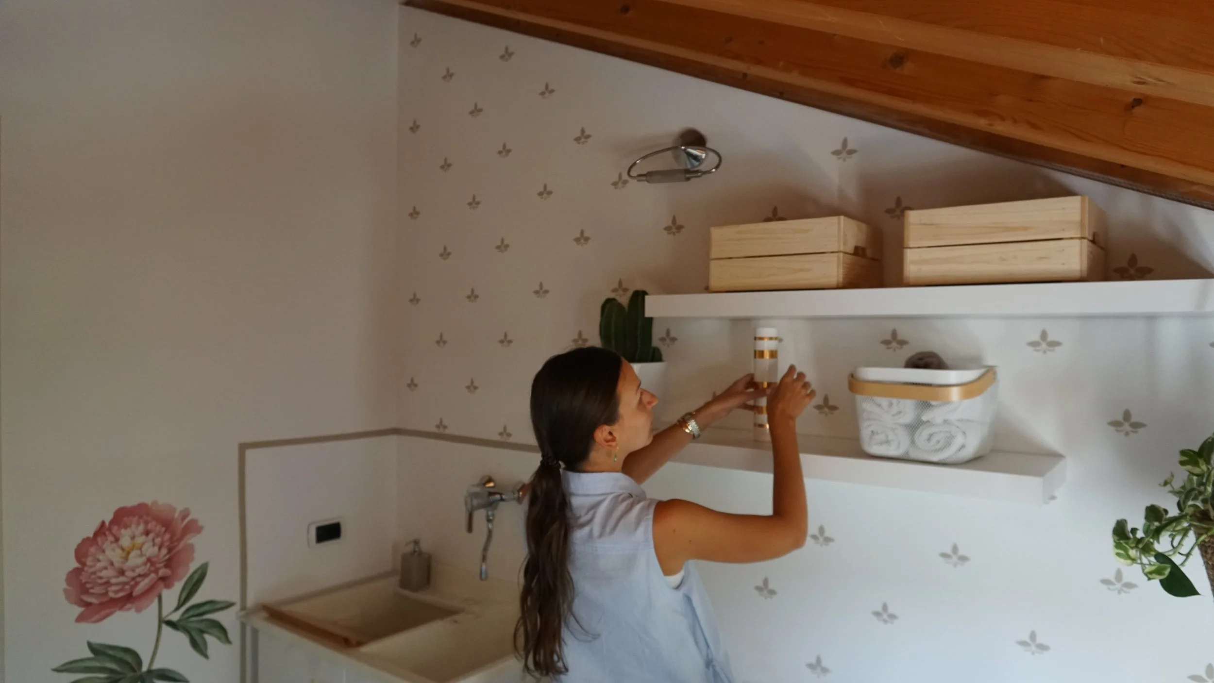 Una donna con capelli lunghi e veste senza maniche appoggia una bottiglia su una mensola bianca, in una stanza con pareti avorio con piccole decorazioni dorate, uno scaffale con scatole di legno, una pianta, un lavandino e un wall sticker con un fiore rosa e foglie verdi.