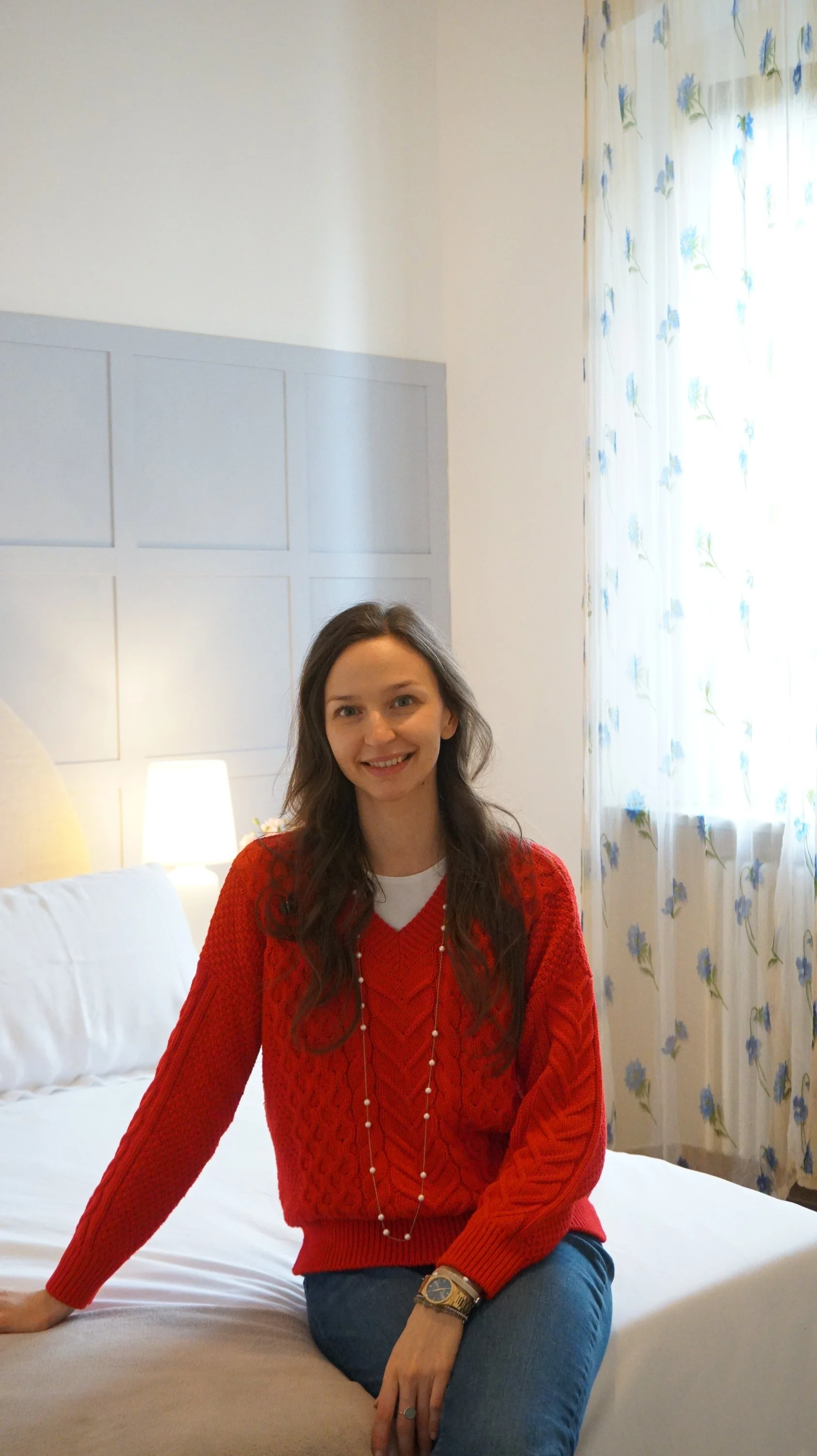 Una donna seduta su un letto in una stanza con tendaggi chiari con motivi floreali, un comodino con lampada acceso dietro di lei.