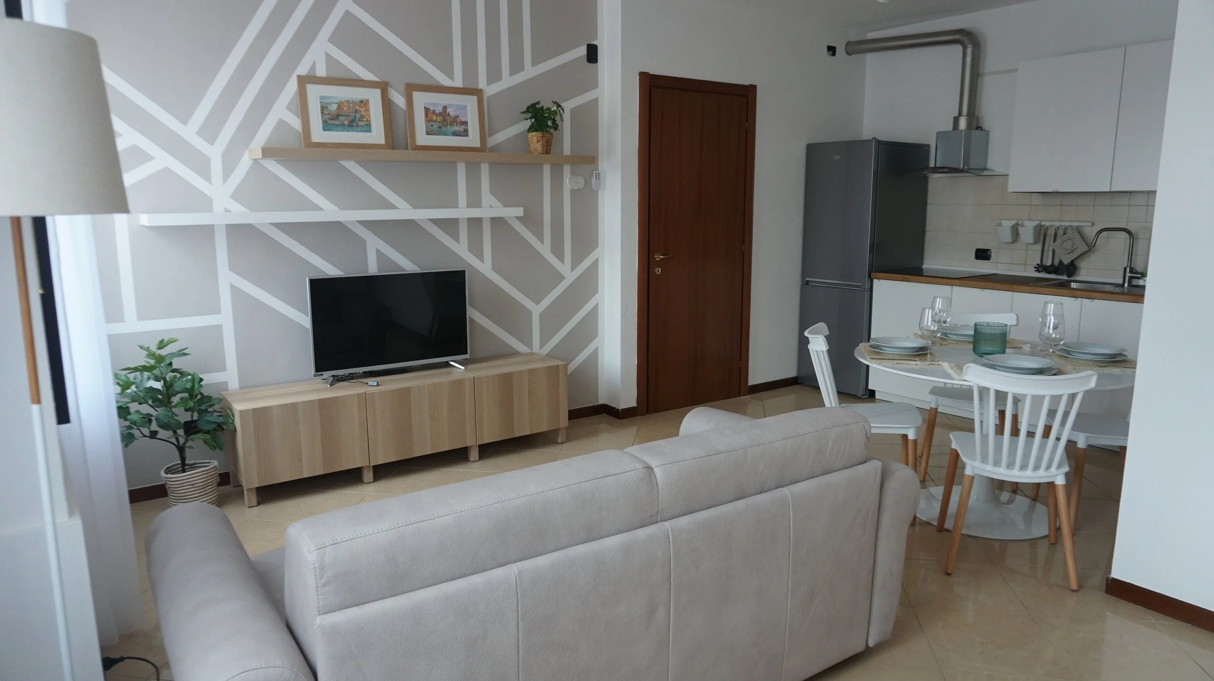 Soggiorno e cucina aperta in un appartamento moderno, con divano beige, televisore su mobile in legno chiaro, parete decorativa con linee bianche, tavolo rotondo con piatti e bicchieri, cucina con frigorifero grande, area lavabo e spazio di lavoro, facili dettagli decorativi.