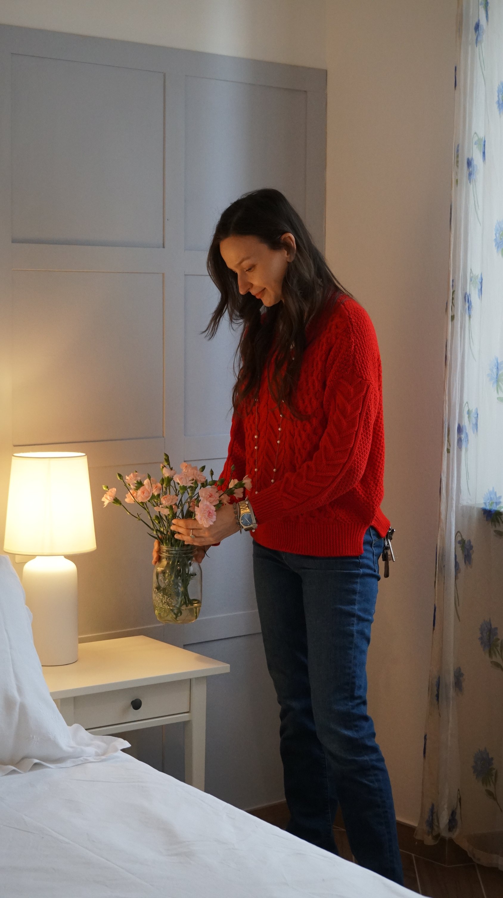 Una donna con maglione rosso che mette dei fiori in un barattolo su una comodino a fianco di un letto in una stanza con pareti chiare e una lampada accesa.