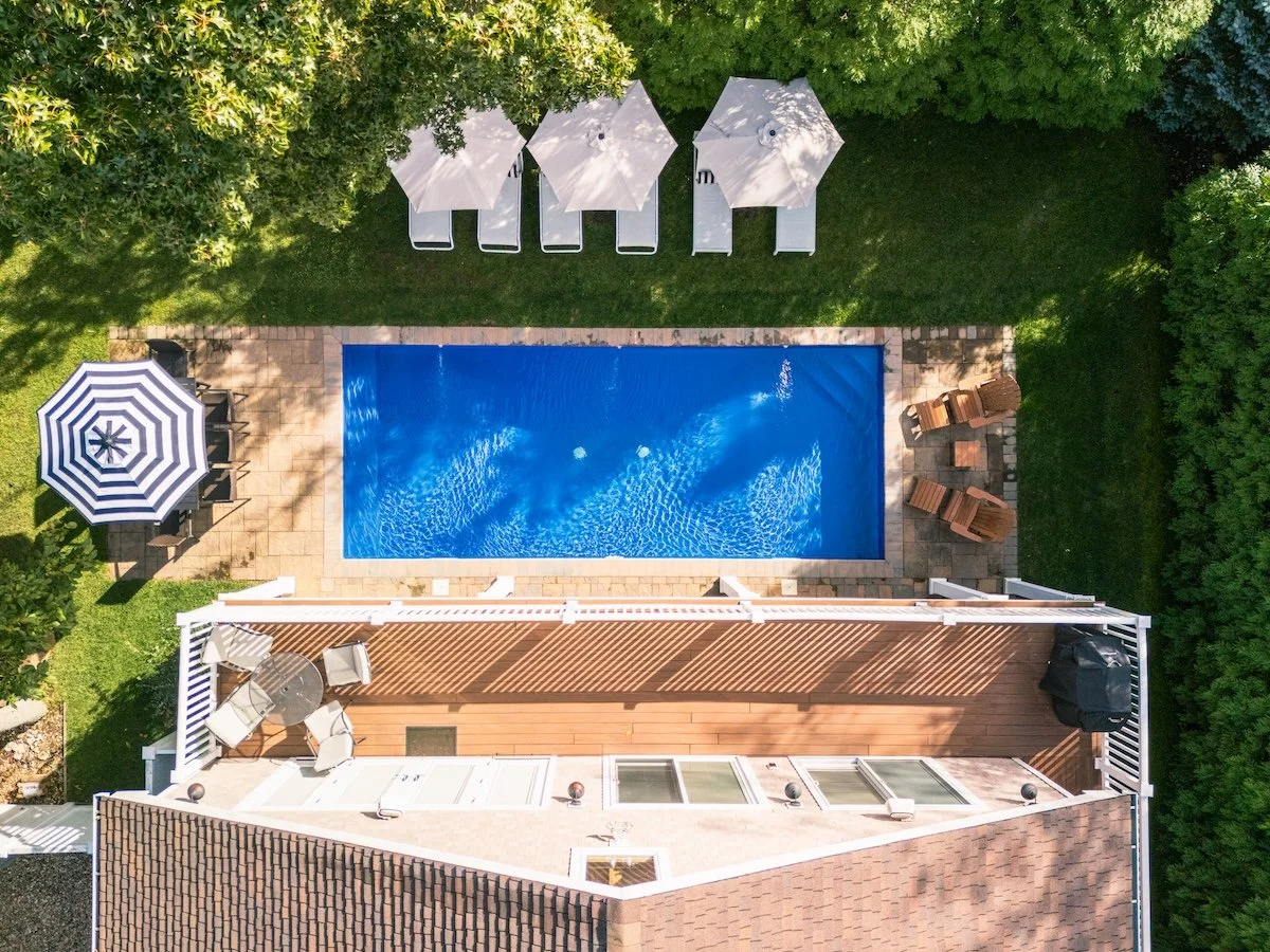 Overhead view of a backyard patio with a swimming pool, surrounded by green trees and grass. There are lounge chairs with umbrellas, patio furniture, and a deck area.
