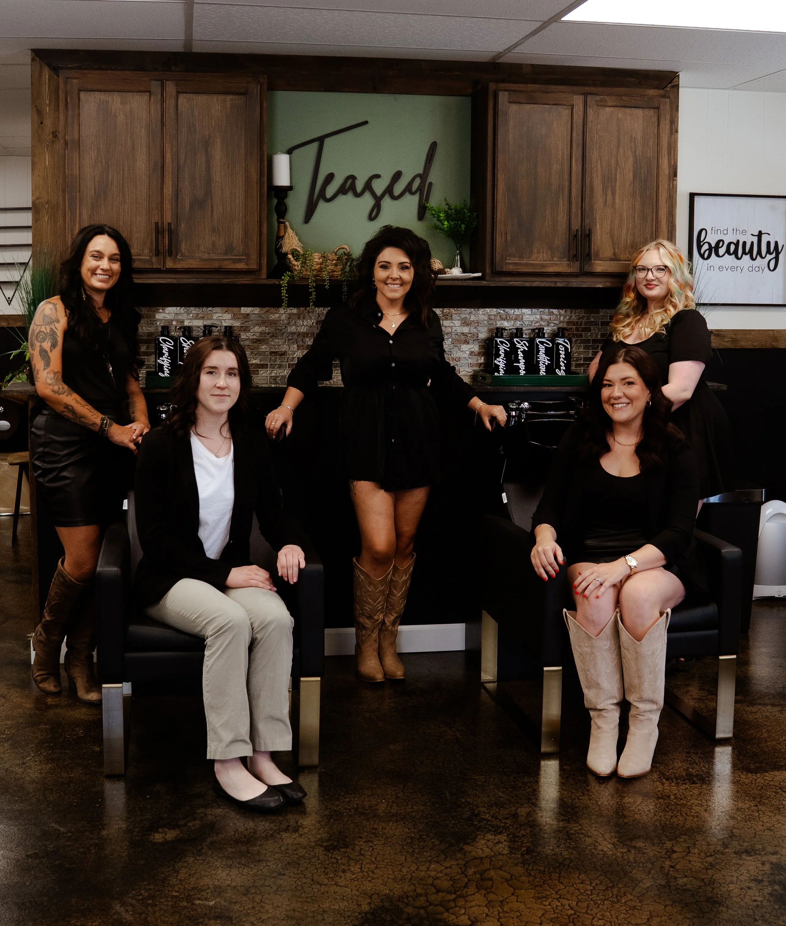A group of five women posing inside a salon with wooden cabinets and decorative signs in the background, dressed in black and neutral clothing.